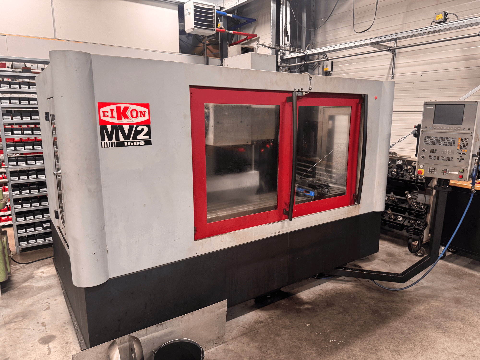 Centre d'usinage vertical Eikon MV2 dans un atelier, vue de face avec des touches de rouge et le panneau de commande visible sur le côté.