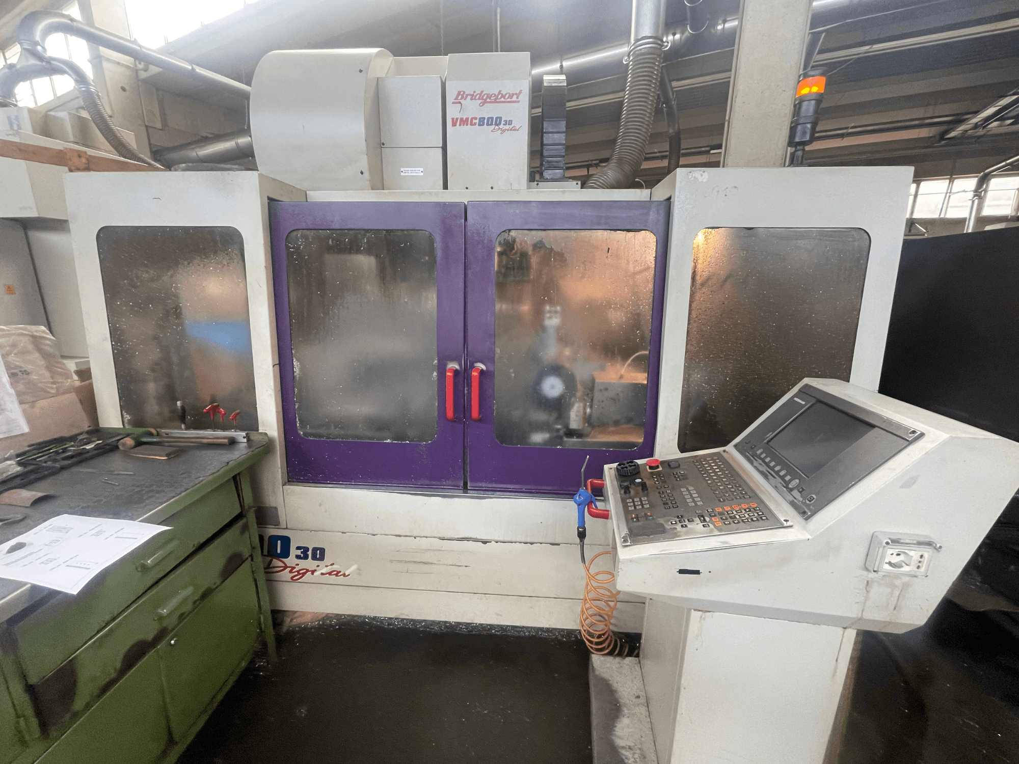 Centre d'usinage vertical Bridgeport VMC 800/30, vue de face, mettant en évidence le panneau de commande et les portes violettes, avec des outils posés sur la surface adjacente.