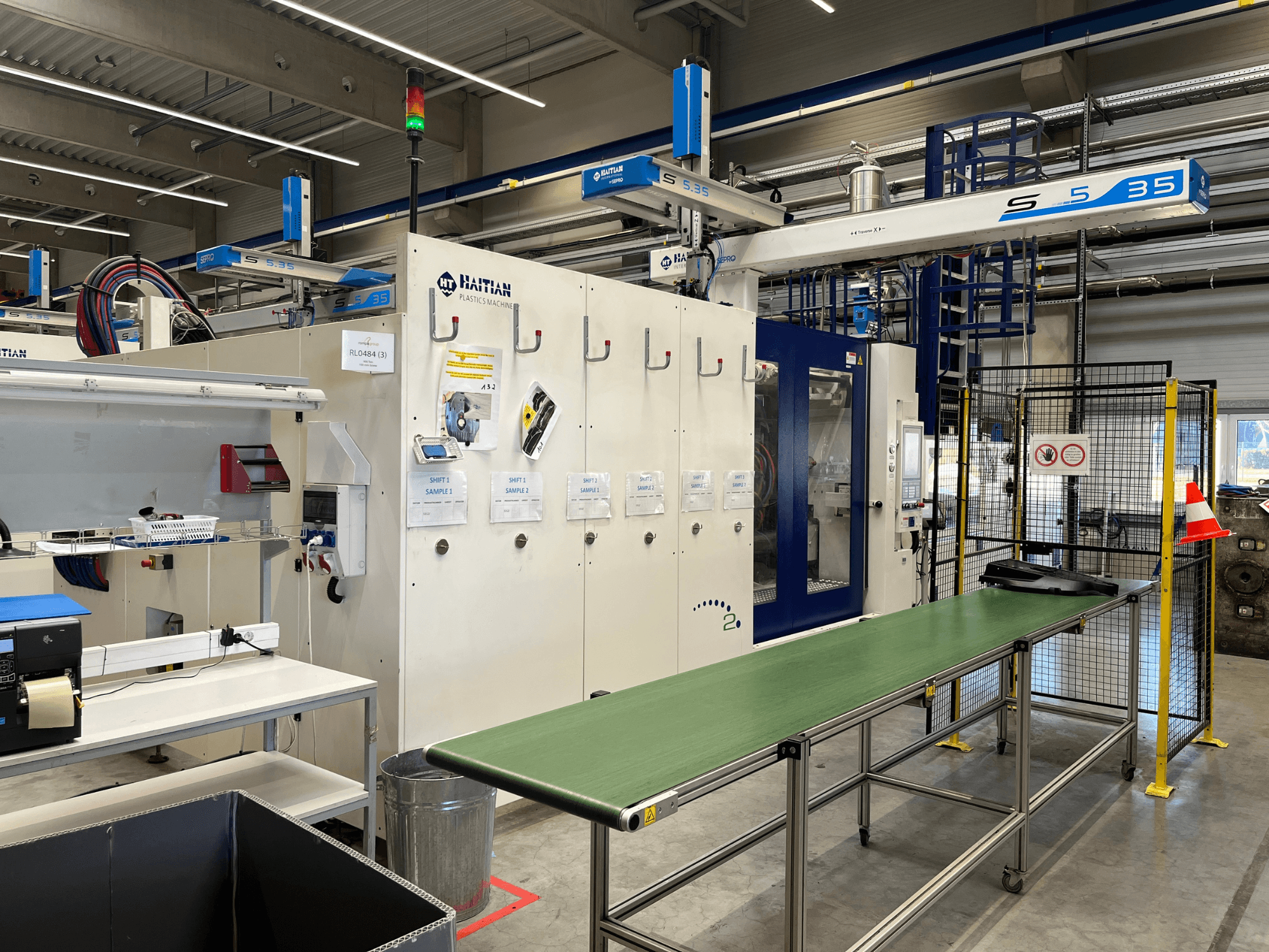 Machine haïtienne S5-35 pour le plastique dans un atelier ; table de convoyage verte, barrières de sécurité et panneau de commande visibles de face.