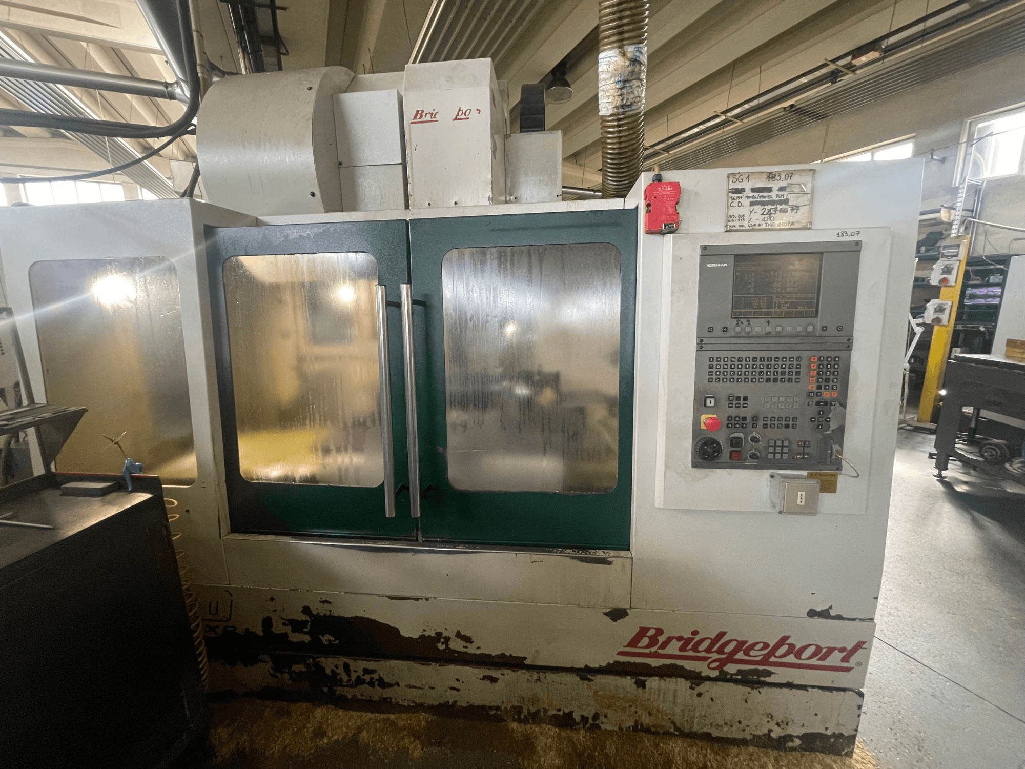Centre d'usinage vertical Bridgeport VMC 1000/30, vue de face avec le panneau de commande, montrant l'extérieur usé et les portes vertes.