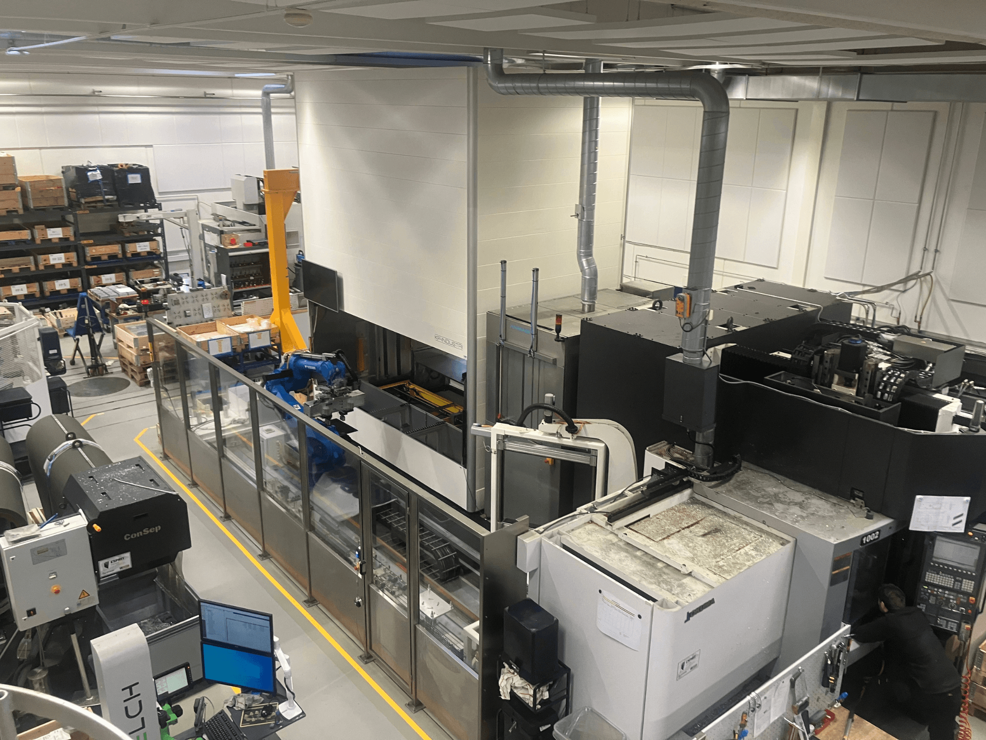 Système d'automatisation Logimat de SSI SCHAFER dans un entrepôt, vue de face ; comprend un bras robotisé, des rayonnages de stockage et des machines.