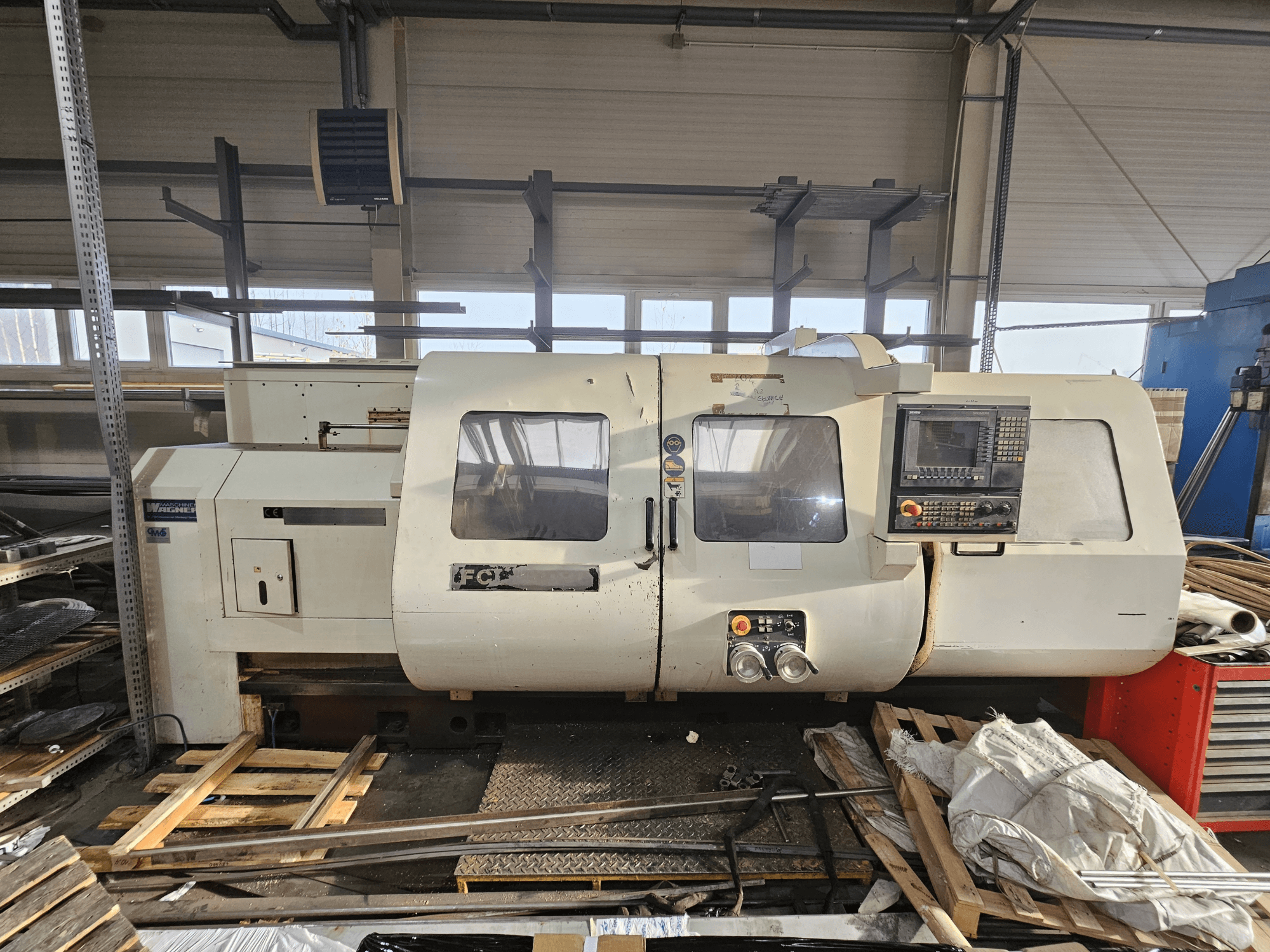 Tour CNC Wabeco, vue latérale, avec panneau de commande, fenêtres et zone de travail ; machines sur palettes en bois dans un atelier.
