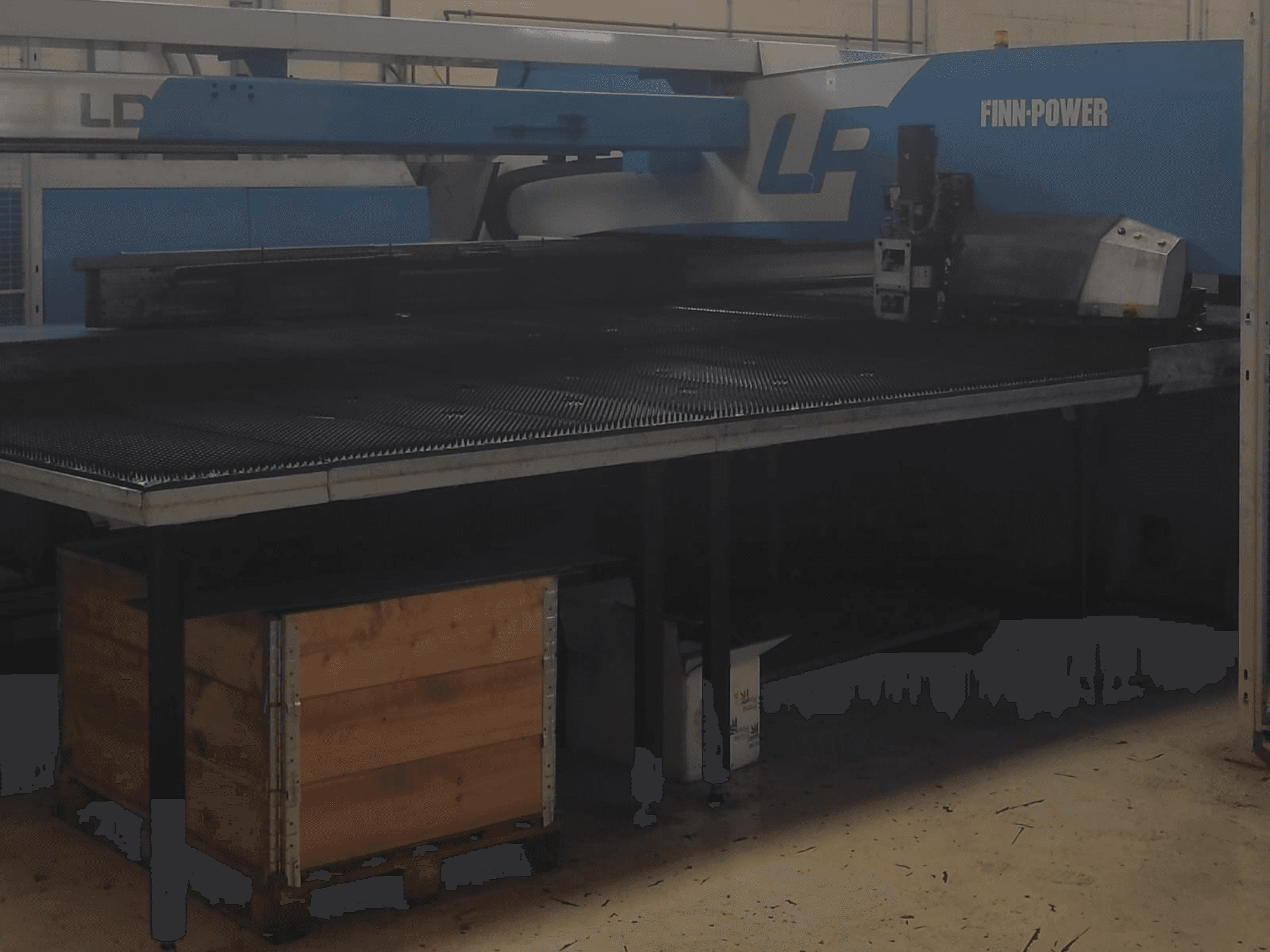 Machine à découper Finn-Power LP dans une usine, vue de face avec une table en métal texturé et une caisse en bois en dessous.