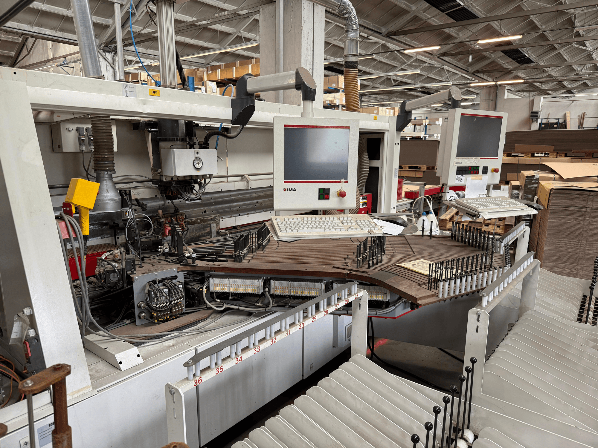 Machine à travailler le bois de précision IMA avec deux moniteurs et panneaux de commande, vue de face montrant le convoyeur et la zone d'outillage.