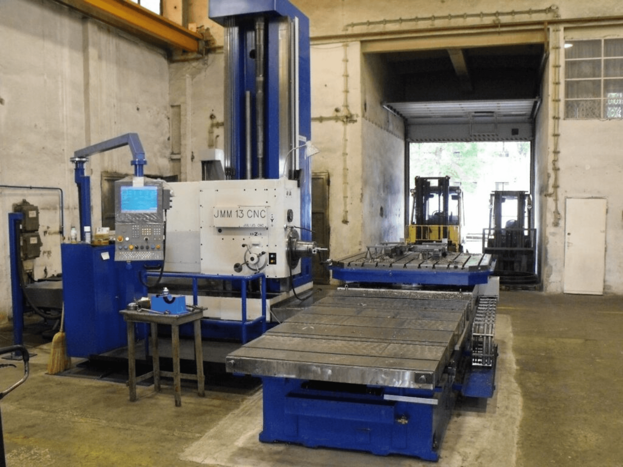 Fraiseuse CNC JMM 13 dans un atelier, vue de face, avec un panneau de commande, une grande table de travail et une toile de fond industrielle.