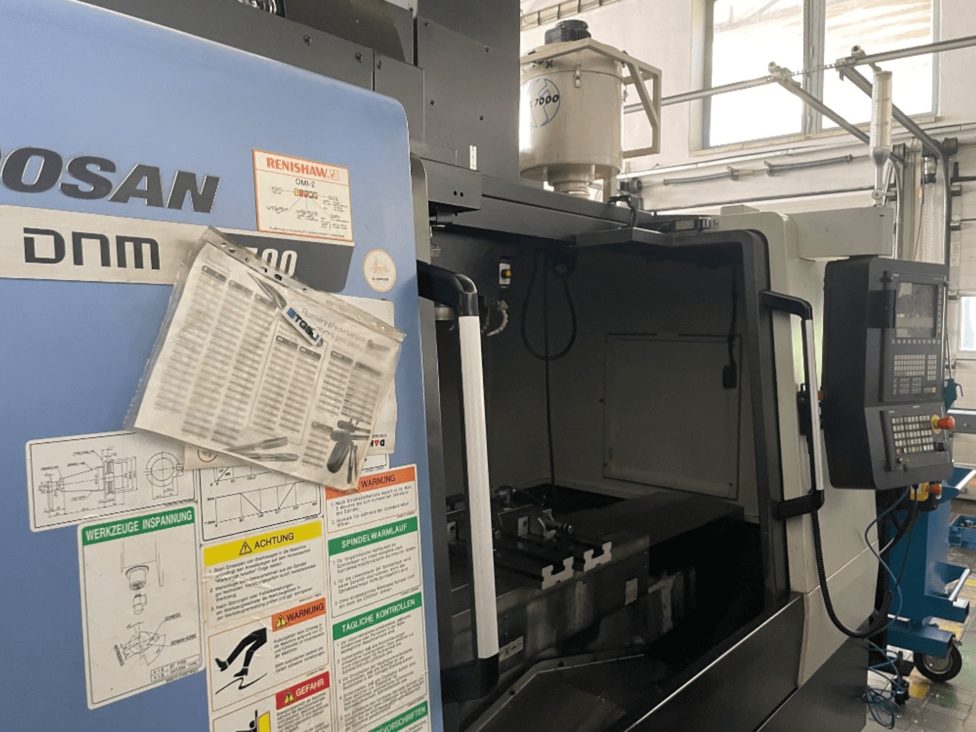 Centre d'usinage vertical DOOSAN DNM 5700, vue de face, montrant le panneau de commande, les étiquettes de sécurité et les détails de la machine.