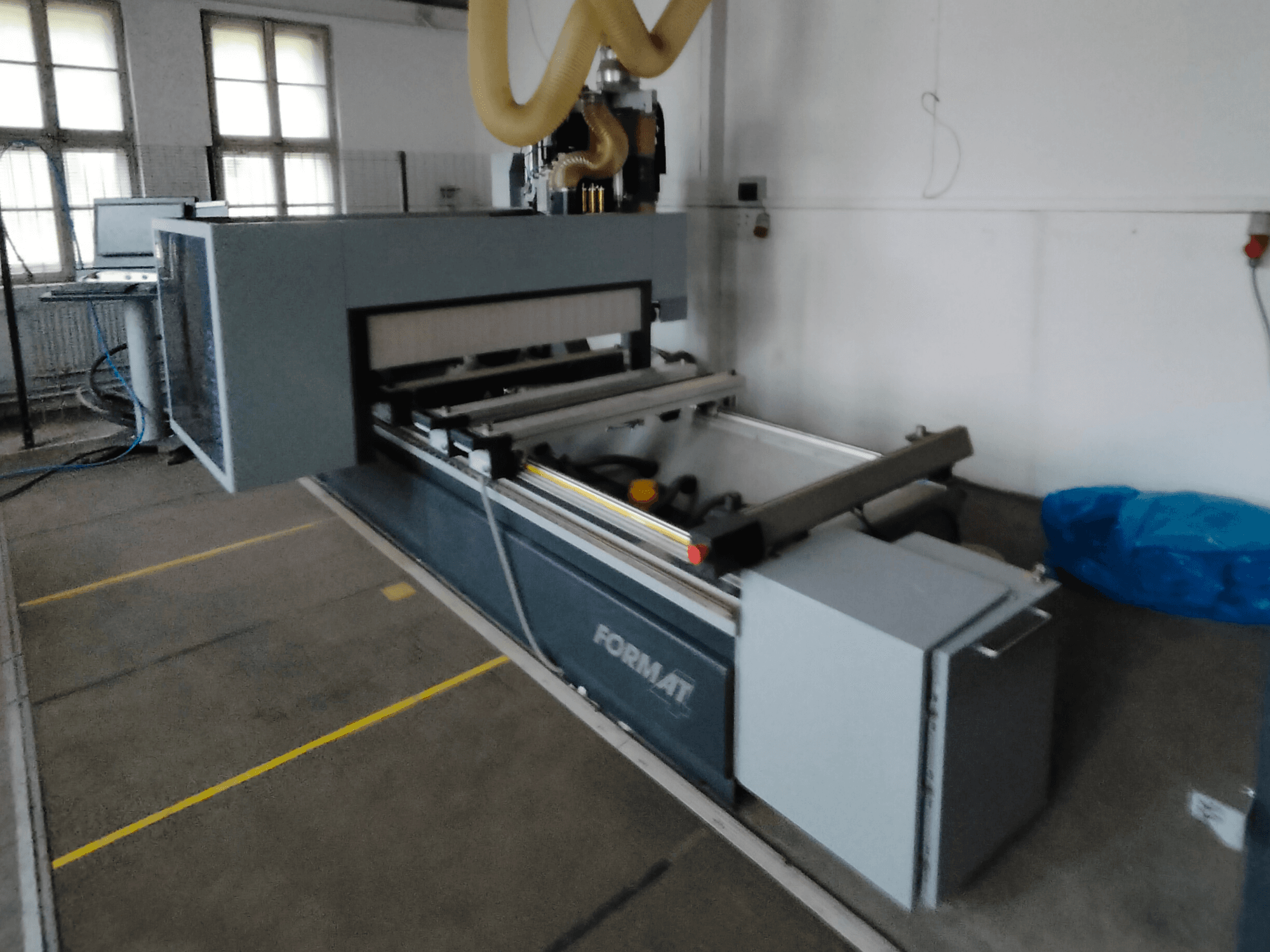 Vue de face d'une machine CNC de format avec un corps gris, comportant des rails de guidage et un panneau de commande visible.