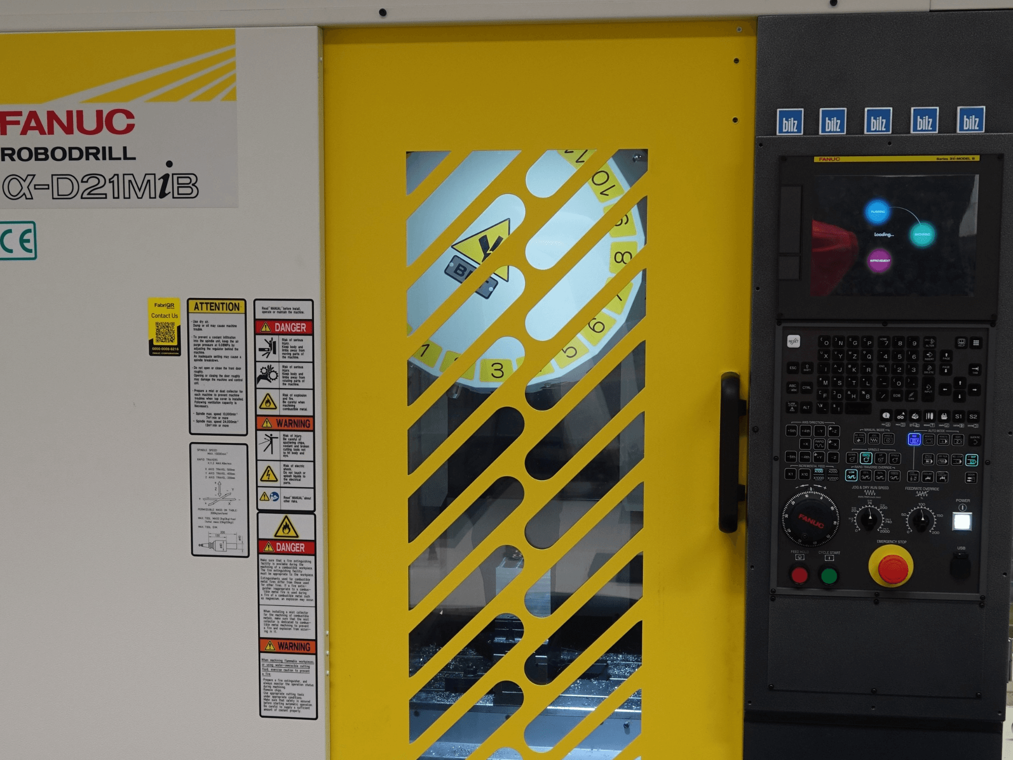 Vue de face du centre d'usinage vertical FANUC α-D21MiB, montrant les avertissements de sécurité, le panneau de commande et la porte de sécurité jaune.