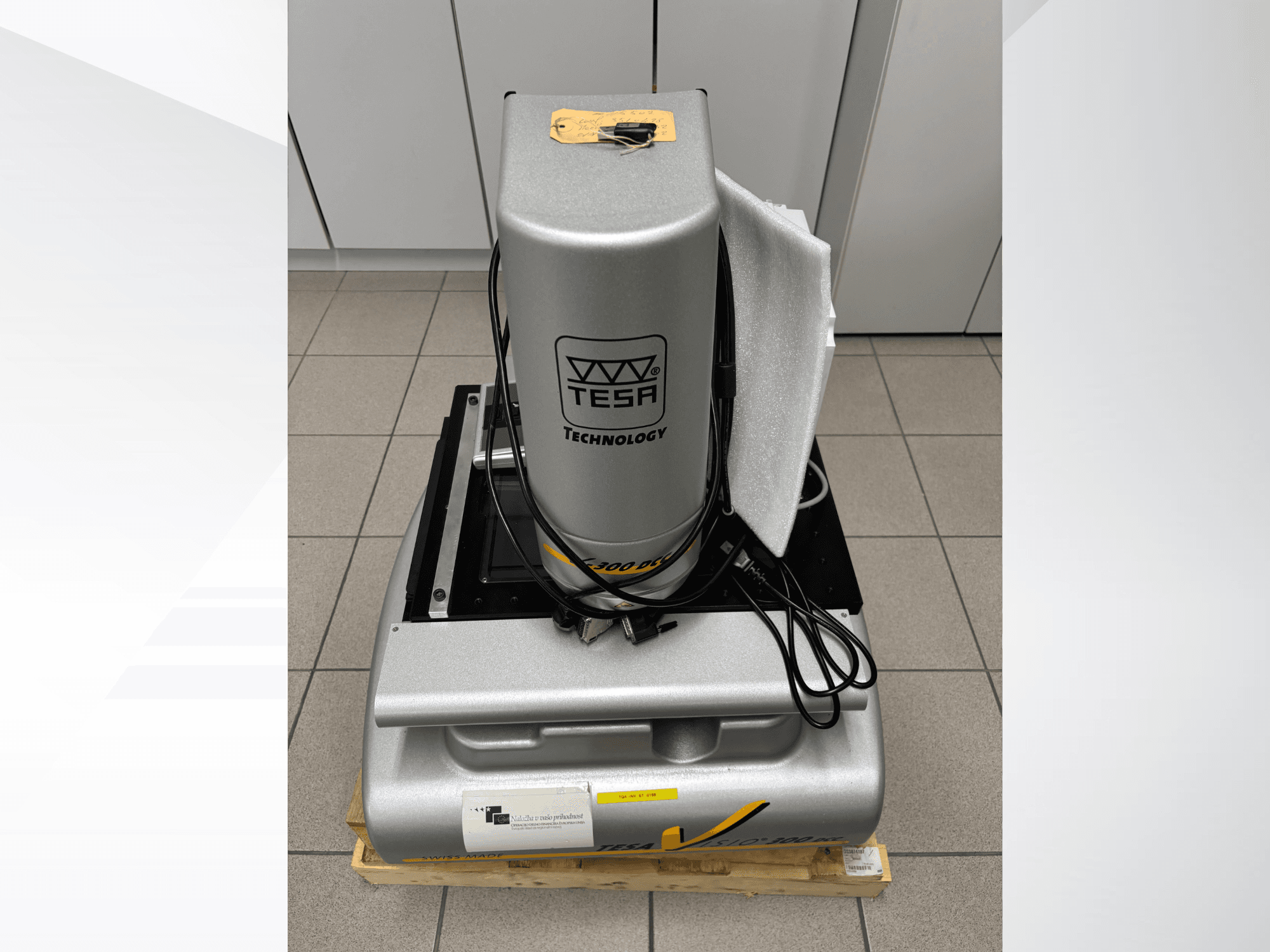 Machine de mesure optique TESA Visio 300 DCC vue de face avec les câbles, sur une palette en bois, et emballage de protection visible.