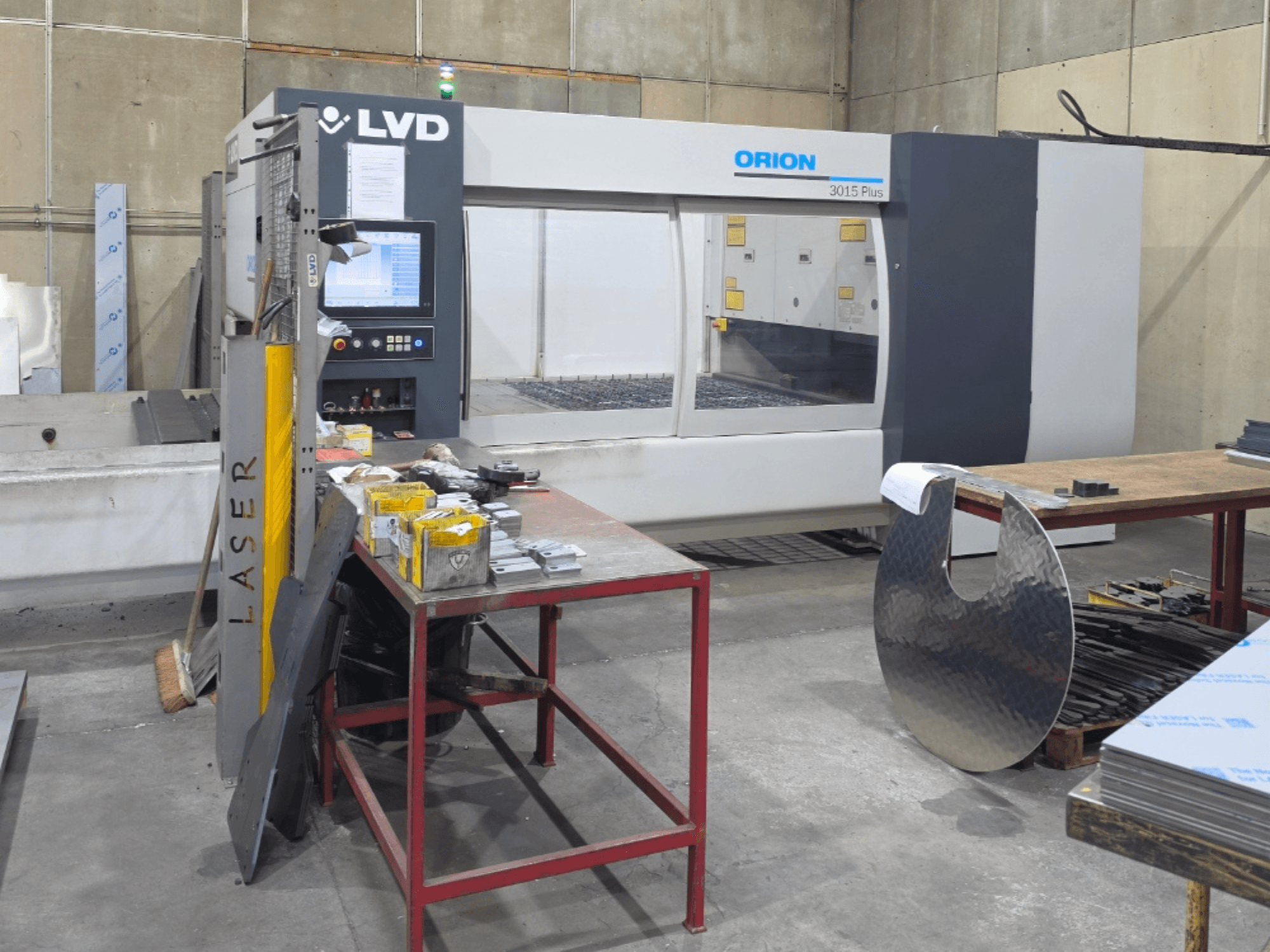 Machine de découpe laser CO₂ LVD Orion 3015 Plus, vue de face avec le panneau de commande ; entourée de pièces métalliques posées sur une table de travail.