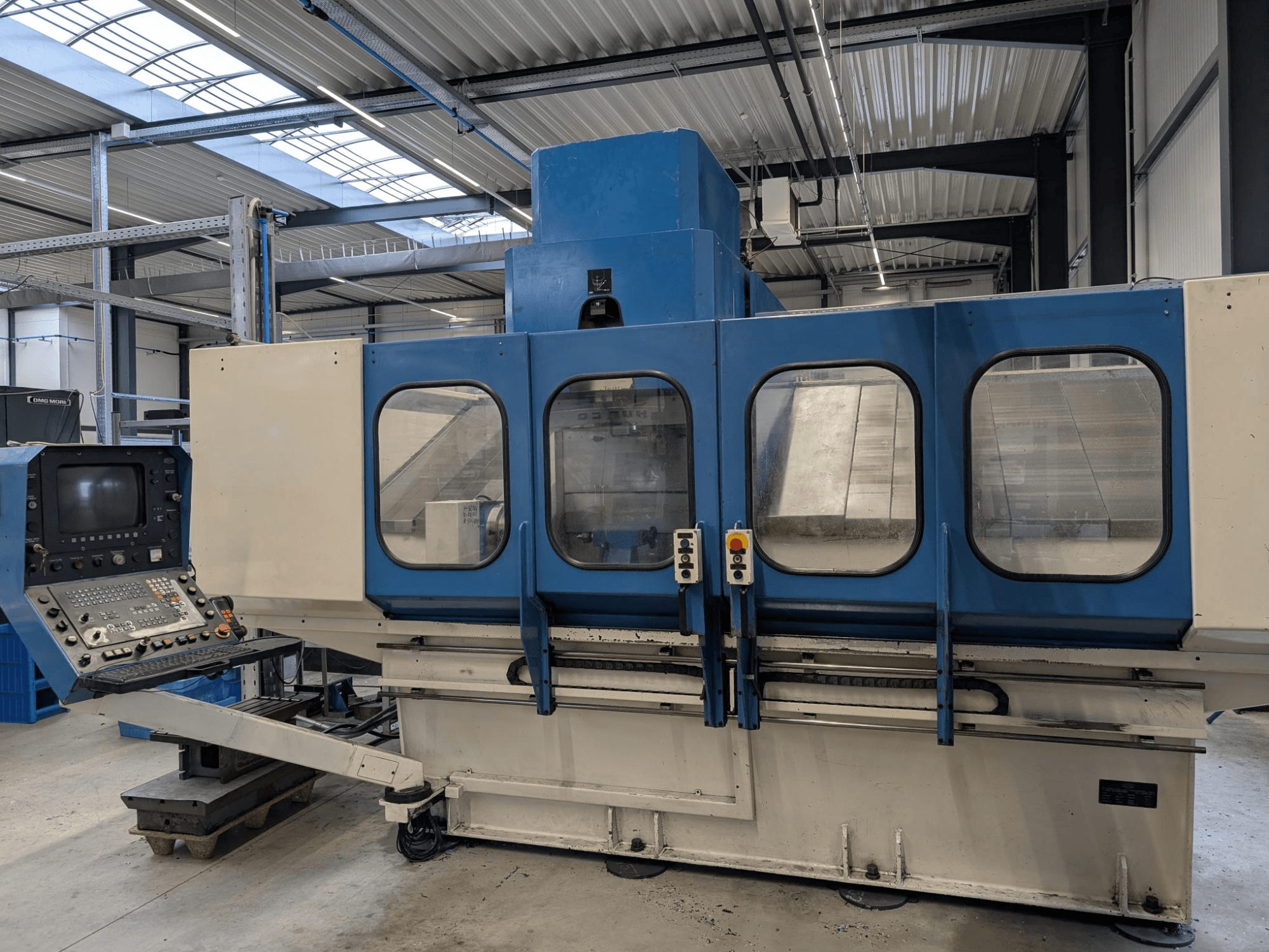 Machine industrielle CNC bleue avec panneau de commande, vue de face. Grandes fenêtres en verre, composants de la machine et boutons visibles.