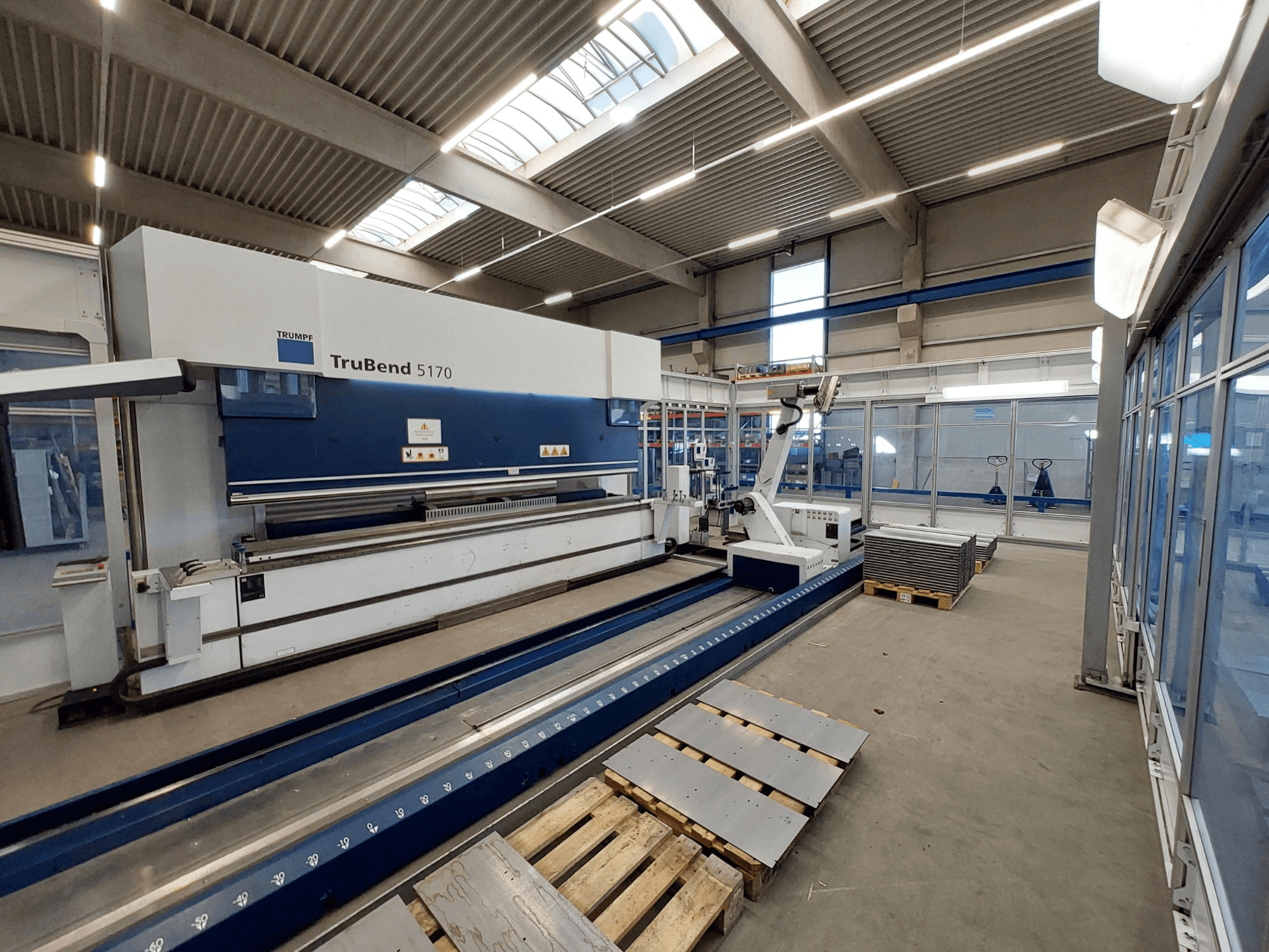 Presse plieuse TRUMPF TruBend 5170 dans un atelier, vue de côté, montrant l'outil de pliage et des tôles empilées sur des palettes.