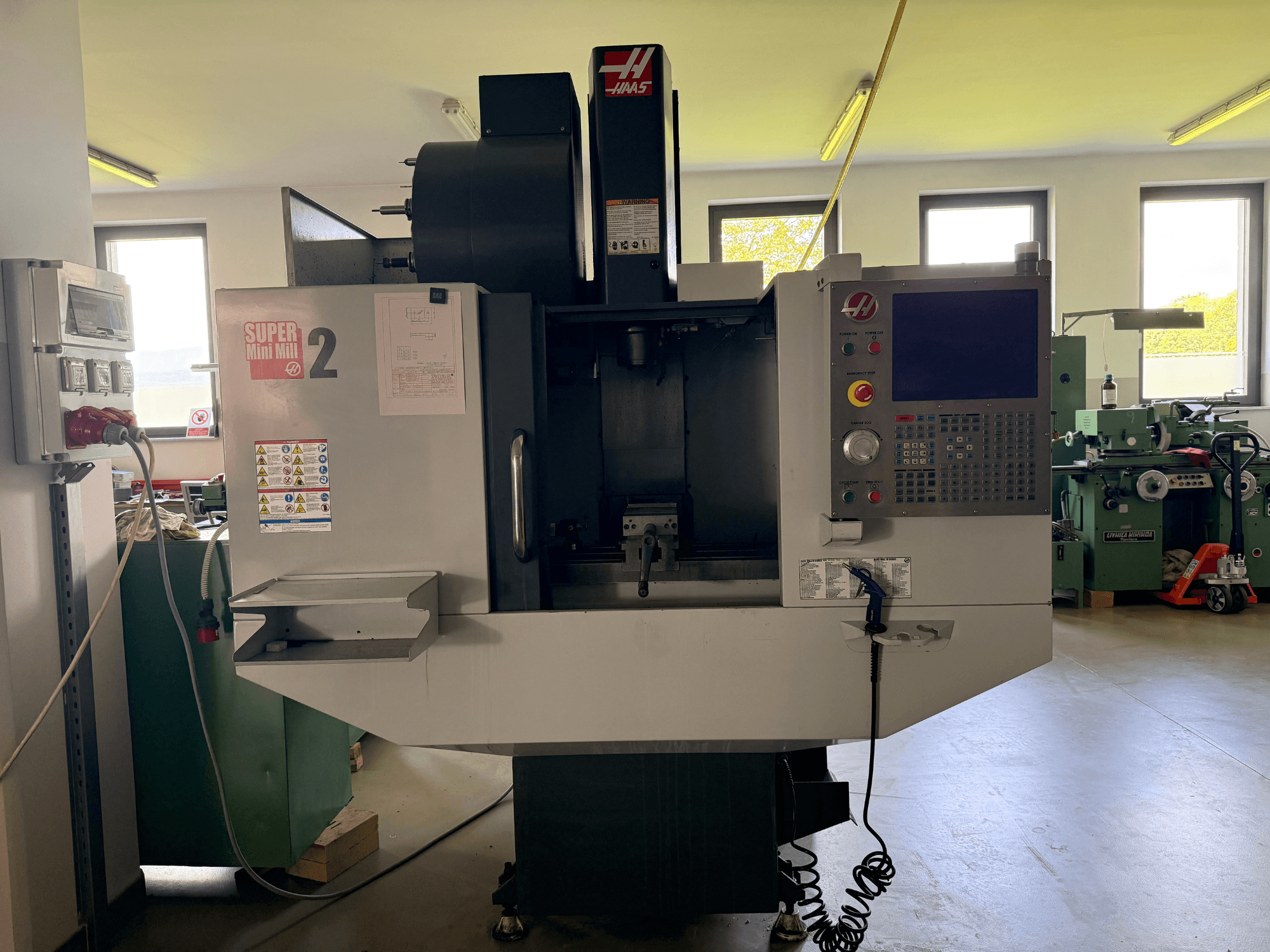 Centre d'usinage vertical Haas Mini Mill 2 en vue de face, présentant le panneau de commande et la zone d'outils avec les étiquettes d'avertissement.