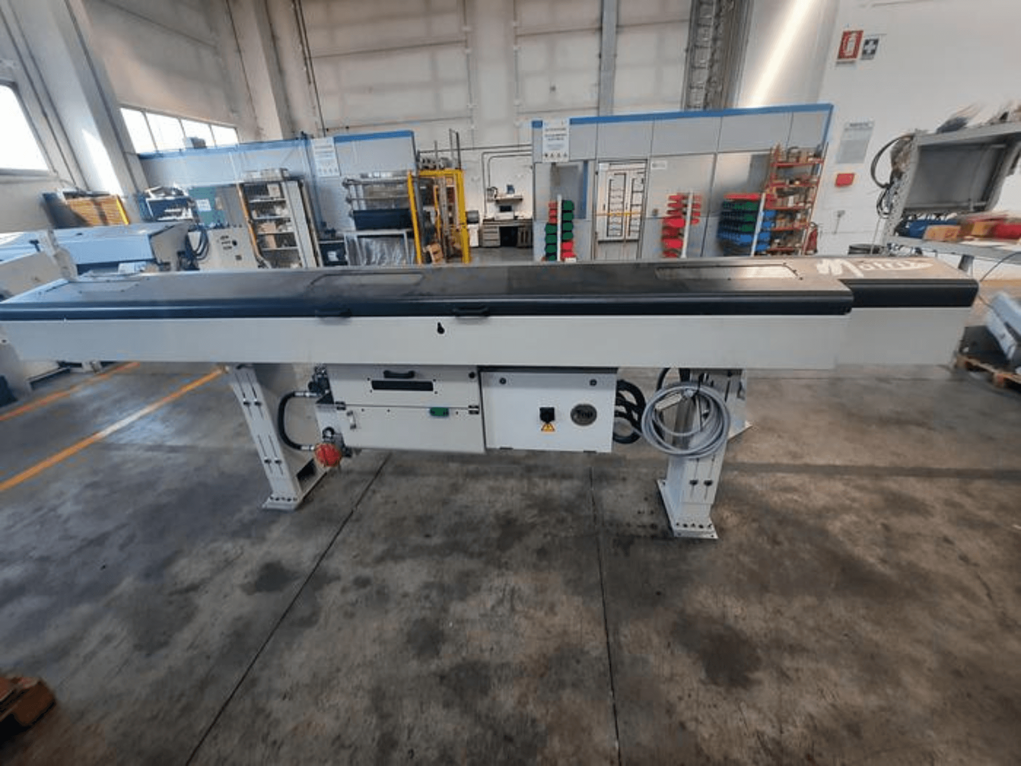 Table de convoyage en métal avec un plateau noir lisse, vue latérale, comportant des boutons de commande et des câbles ; le logo de la marque est partiellement visible.