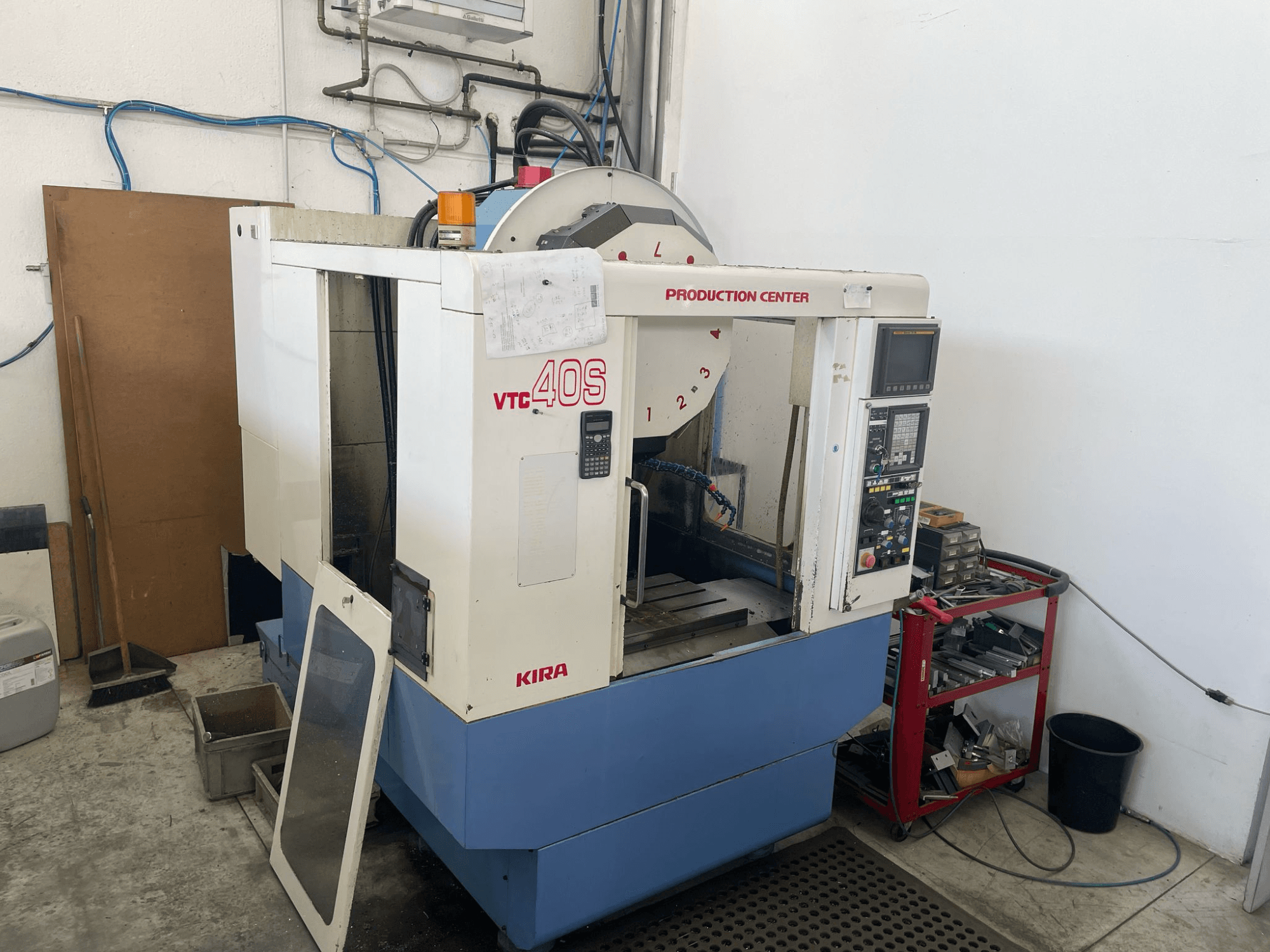 Centre d'usinage KIRA VTC 40S en vue latérale, avec le panneau de commande, la porte ouverte et les outils sur un chariot de stockage à proximité.