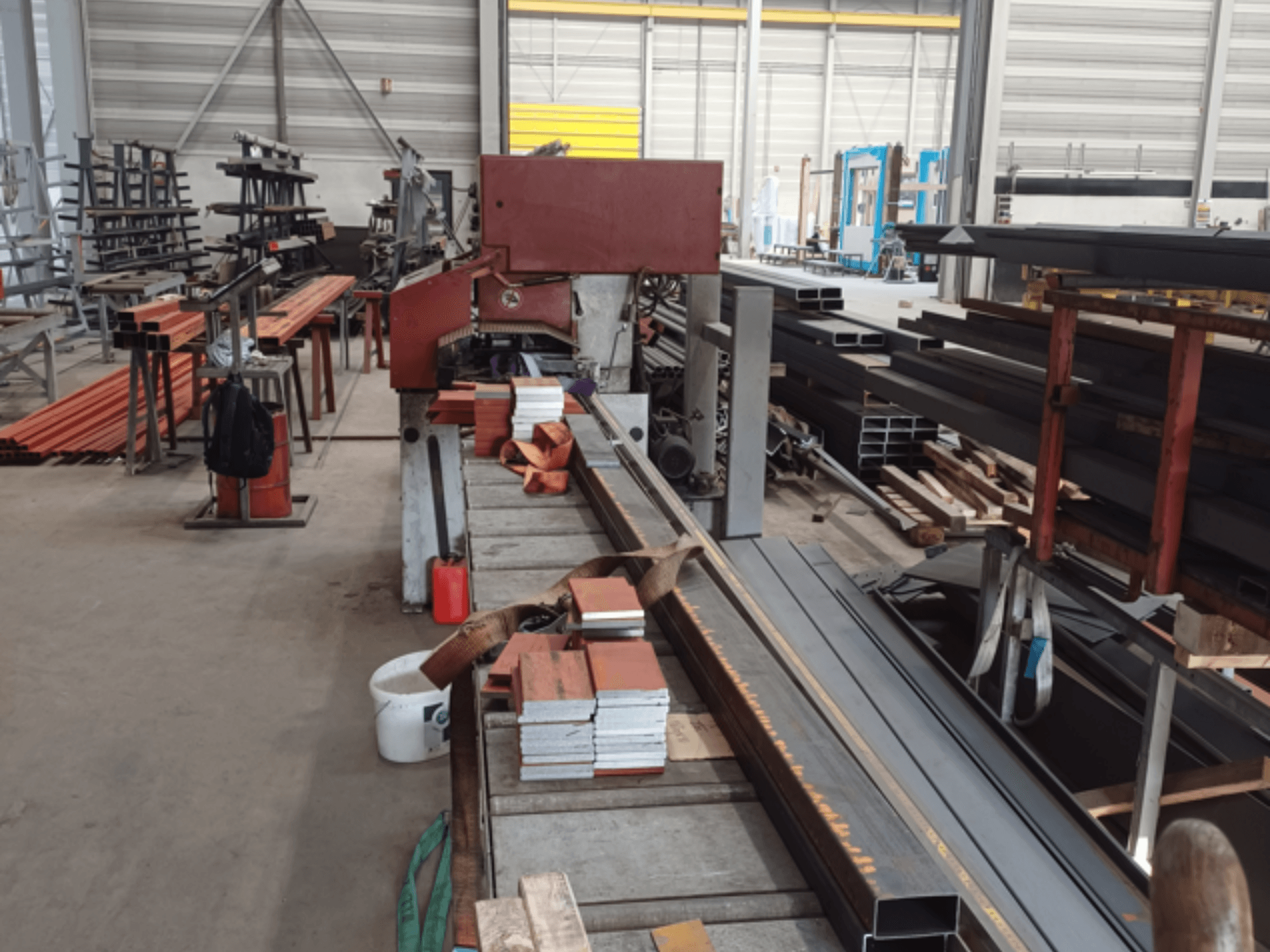 Atelier de fabrication de divers profilés métalliques, avec une scie rouge au premier plan et des matériaux empilés.