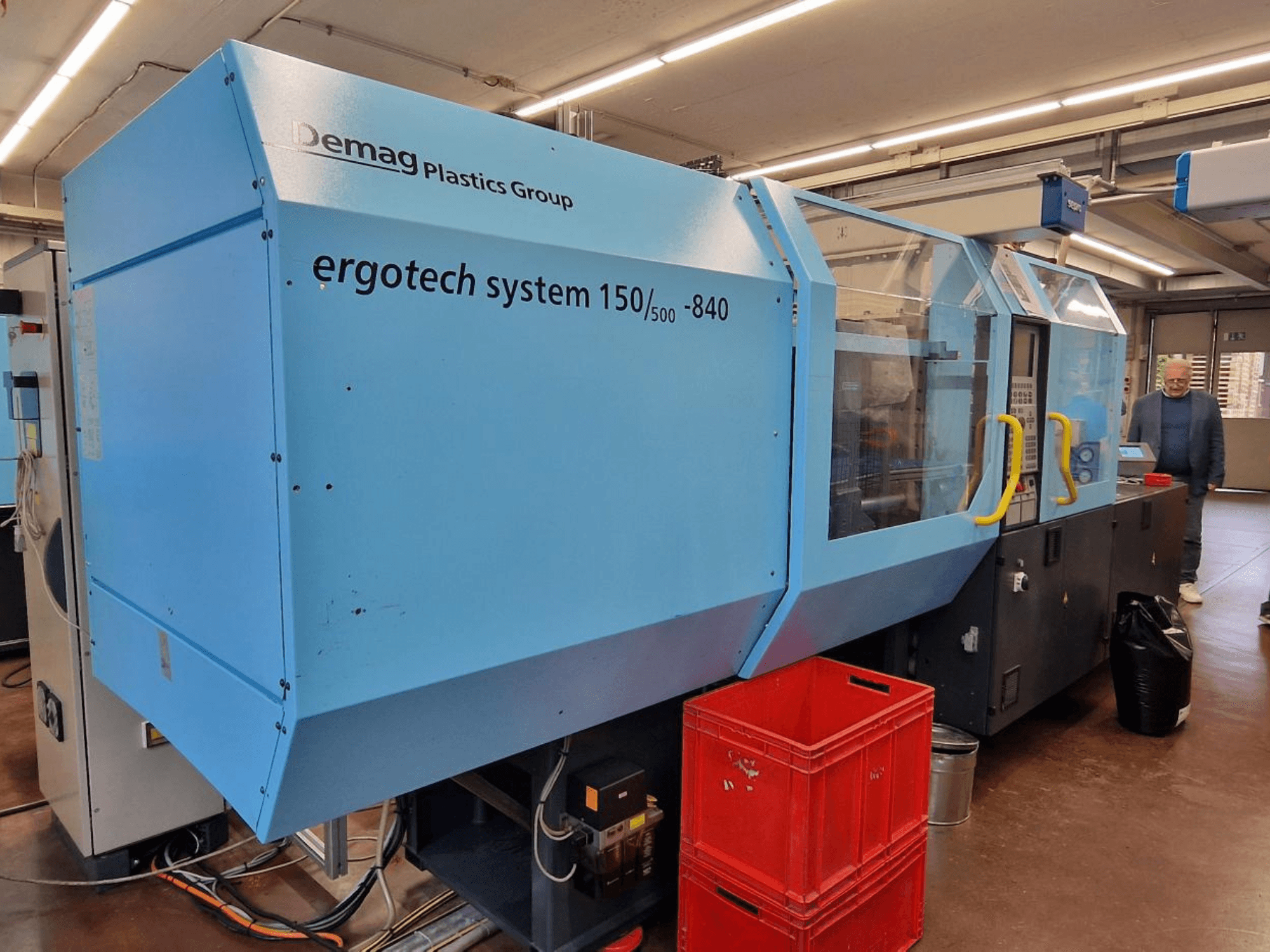 Machine de moulage par injection Demag Ergotech System 150/500-840 en bleu, vue de côté, avec des caisses et un opérateur en arrière-plan.