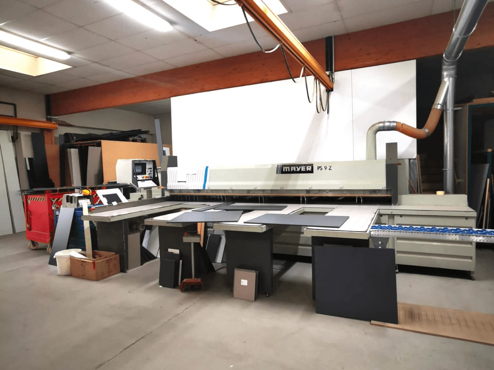 Machine à découper MAYER PS9 Z dans un atelier, vue de face, avec divers matériaux et un système de convoyage visible.