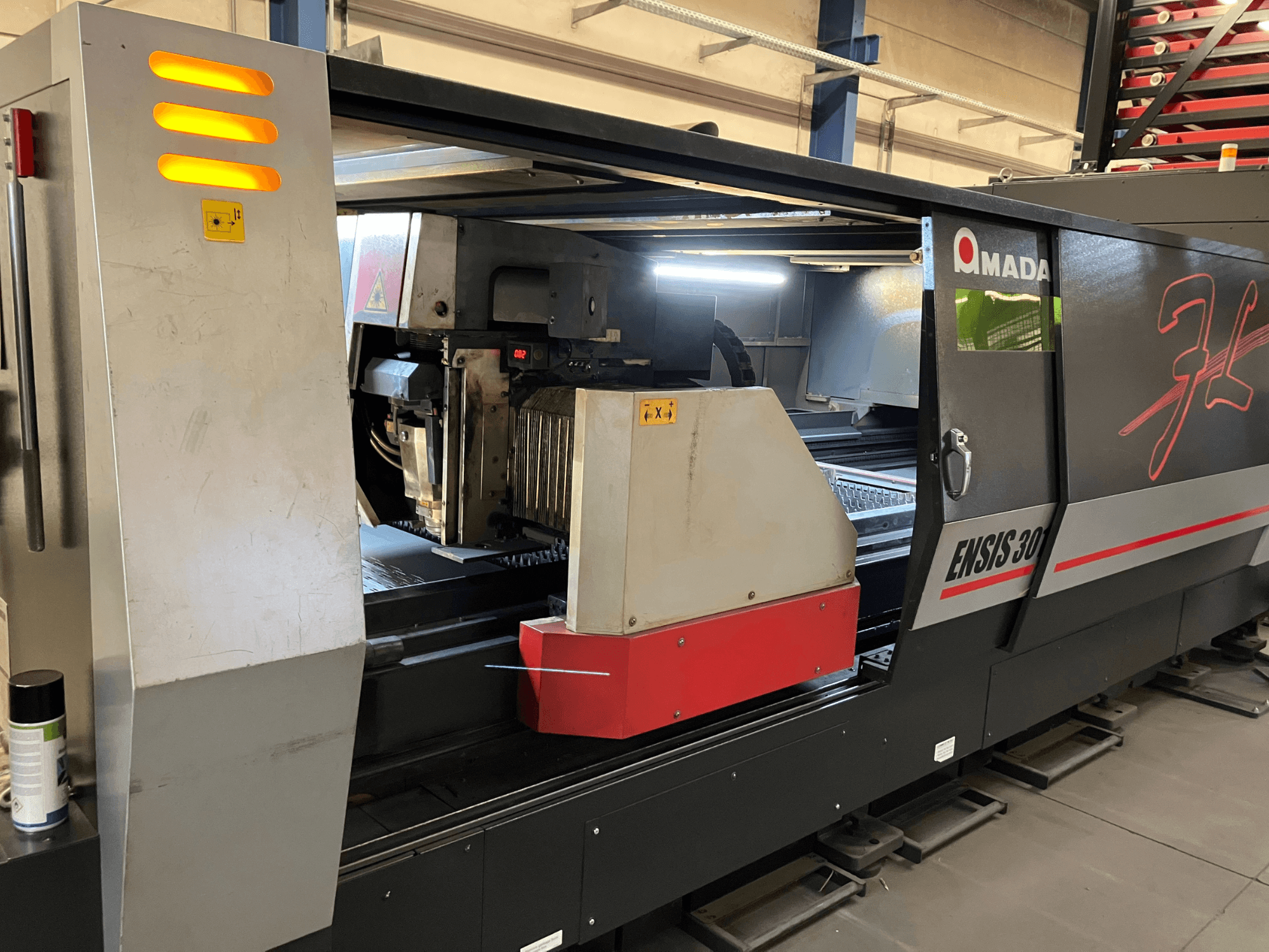 Machine de découpe laser à fibre AMADA ENSIS 3015 AJ, vue de face, montrant la tête de découpe et les commandes à l'intérieur d'un cadre métallique.