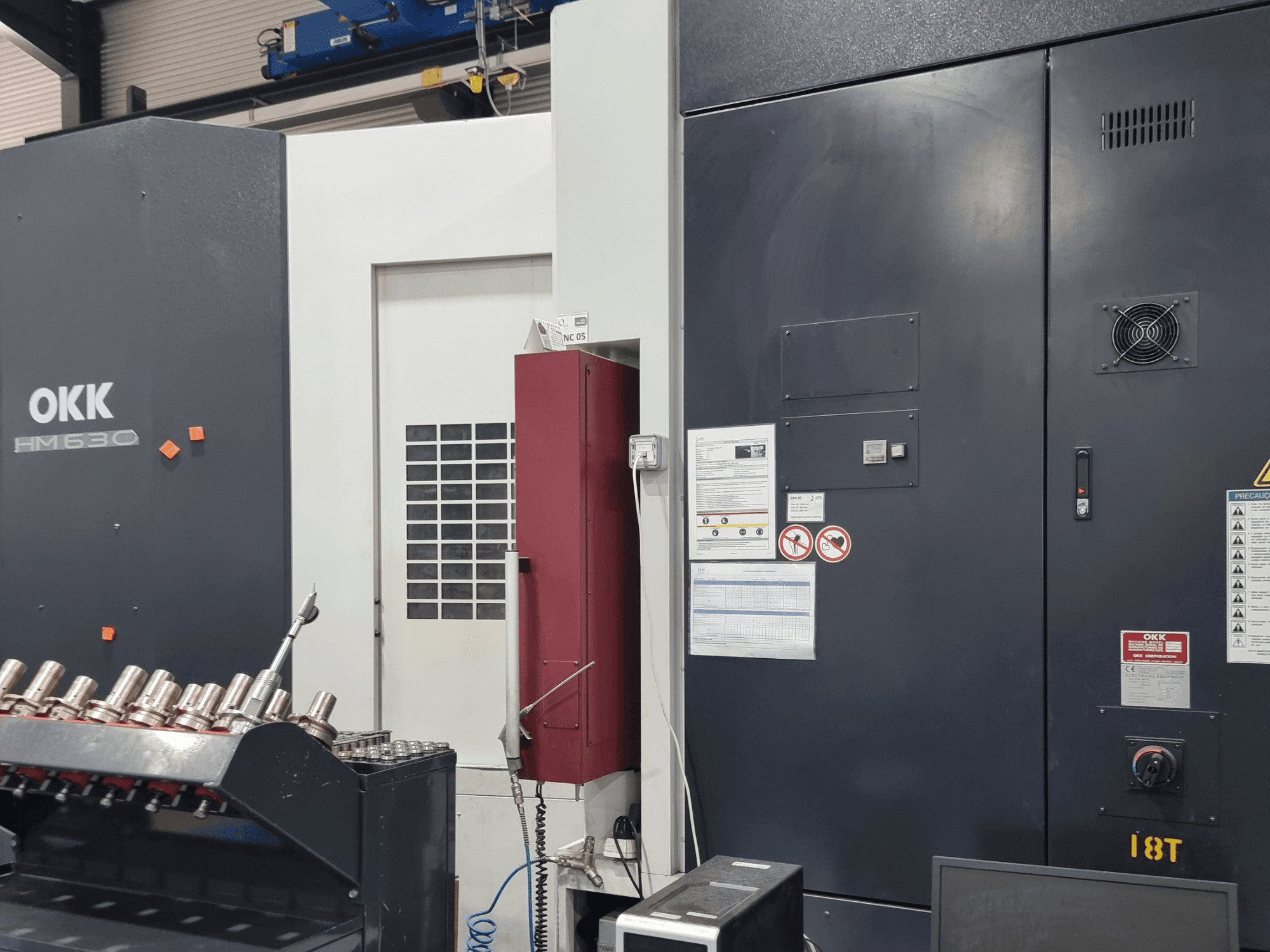 Centre d'usinage horizontal OKK HM630, vue de face montrant le panneau de commande, les consignes de sécurité et le rangement des outils sur le côté.