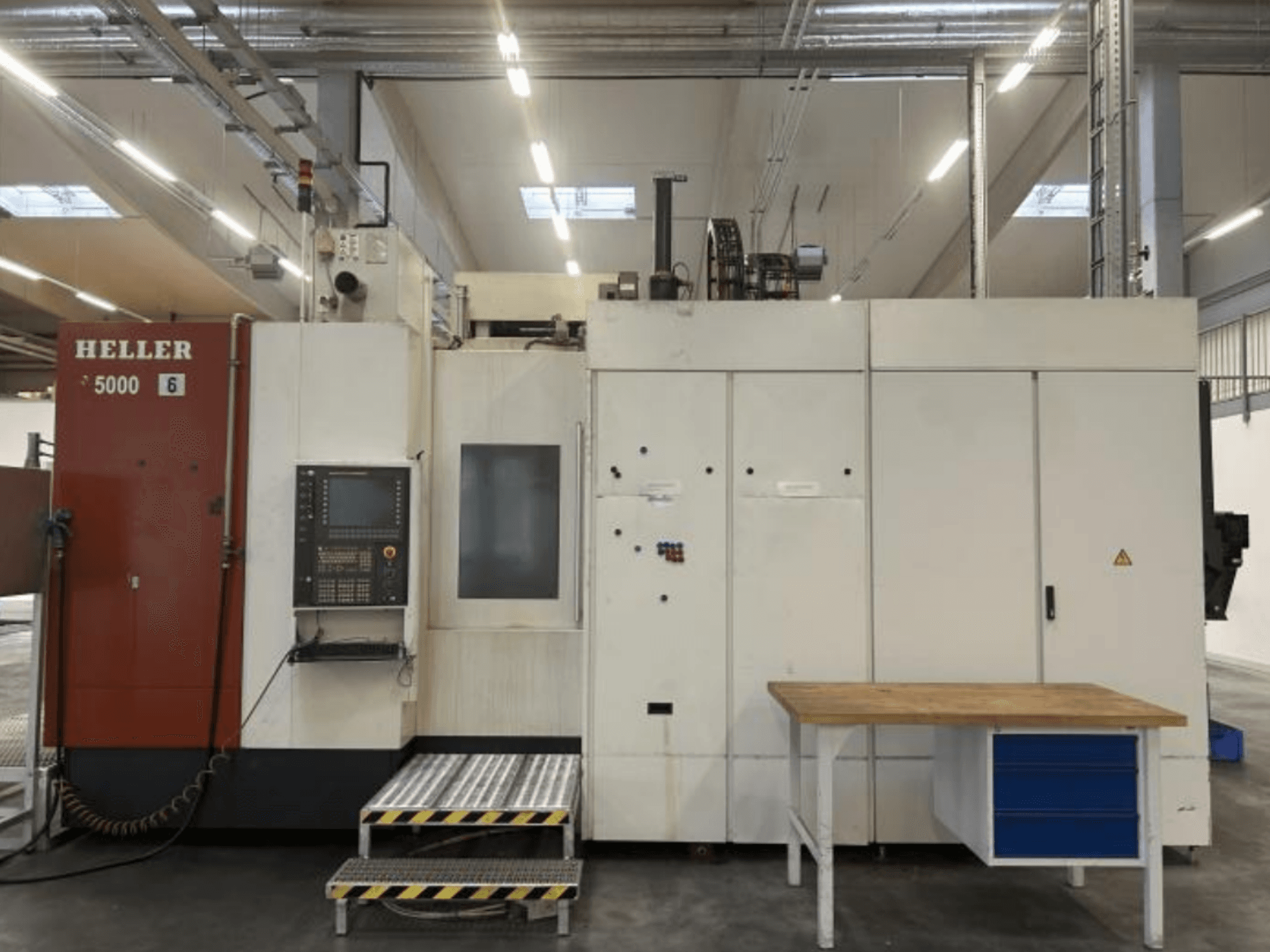 Vue de face du centre d'usinage horizontal HELLER H5000 avec son panneau de commande, sa plate-forme surélevée et sa table de travail adjacente.