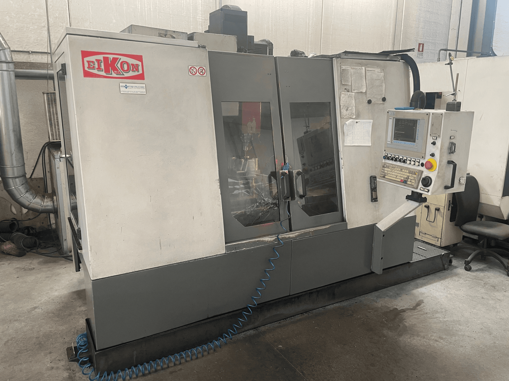 Machine CNC EIKON vue de face, avec un panneau de commande, de grandes portes vitrées et un tuyau d'air bleu au sol.