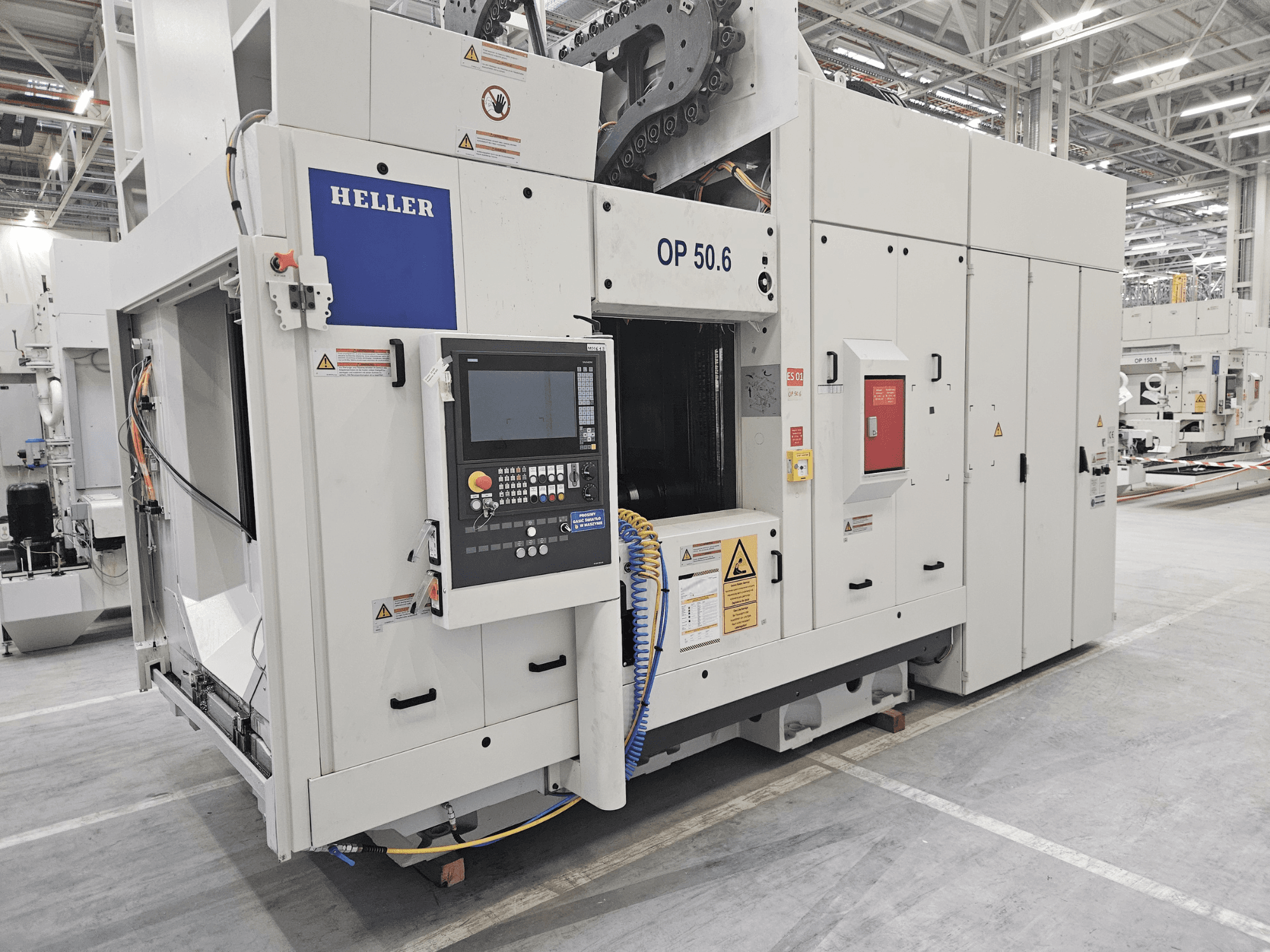 Centre d'usinage Heller OP 50.6, vue de face, avec un panneau de commande, des étiquettes d'avertissement et des tuyaux visibles en usine.