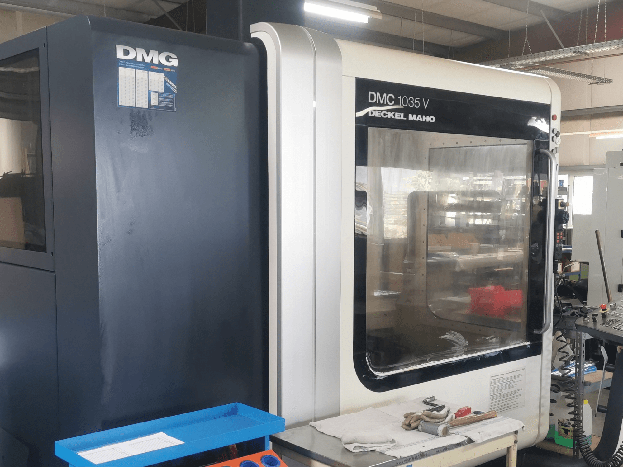 Fraiseuse DMG DMC 1035 V dans un atelier, vue de face avec un plateau d'outils bleu et des gants sur la surface.