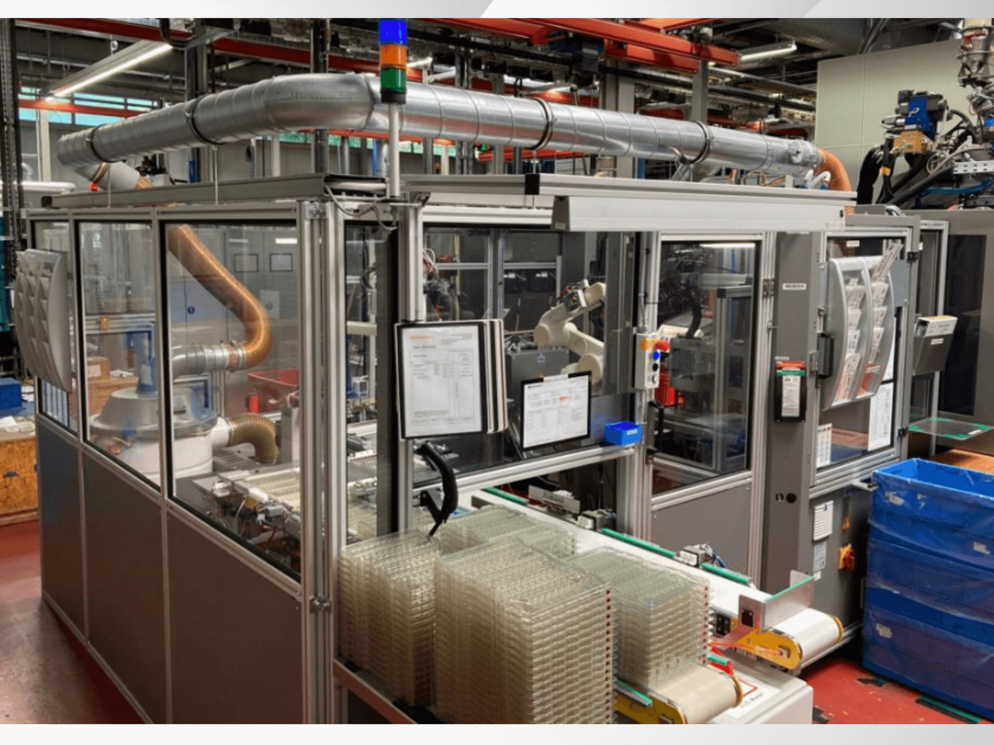 Machine d'assemblage automatisée avec bras robotisé, panneaux latéraux transparents et conteneurs empilés d'articles transparents, vue de face.