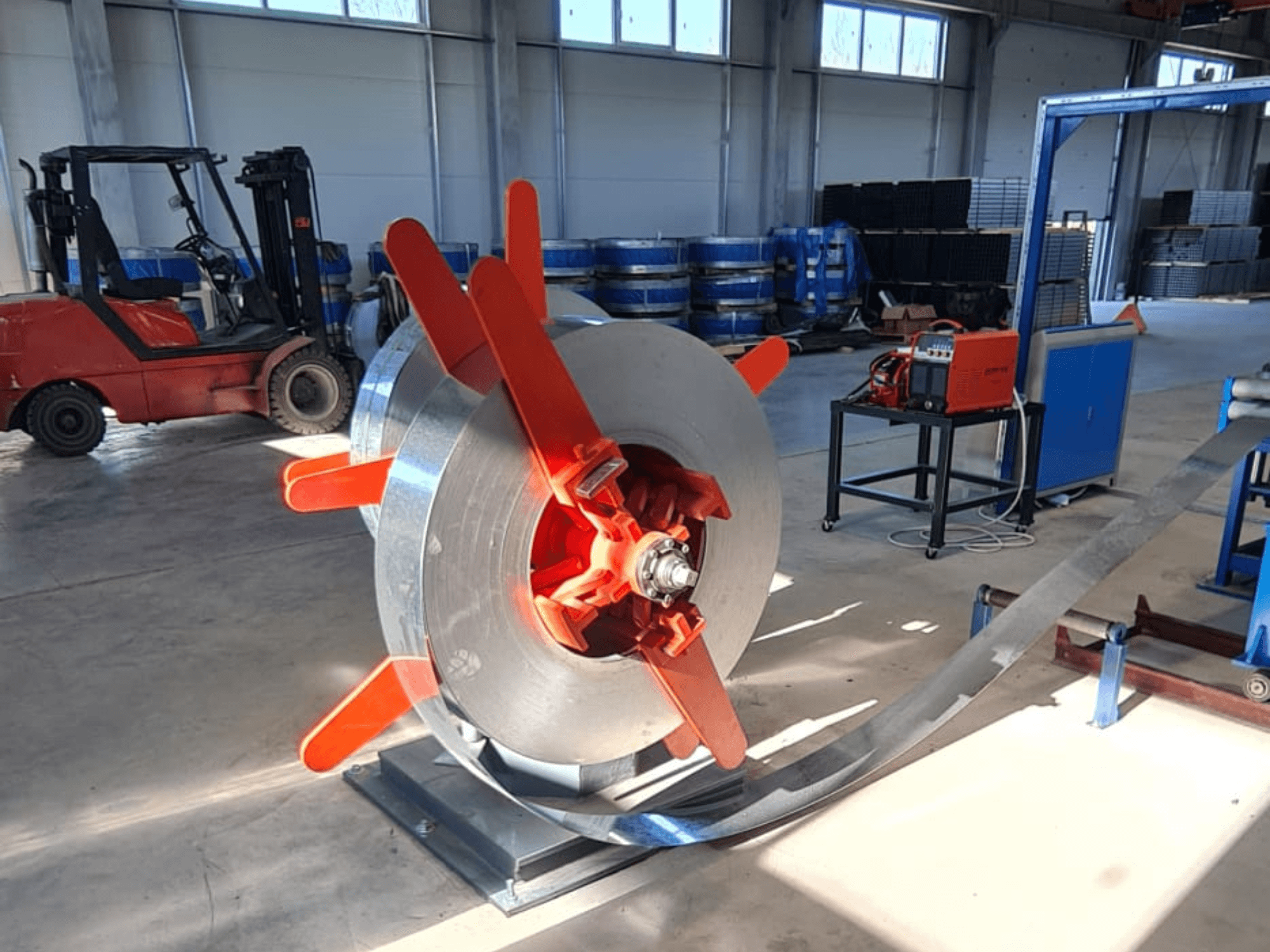 Bobine de métal avec poignées rouges sur un support, vue d'un angle. Arrière-plan industriel avec des machines et un chariot élévateur.