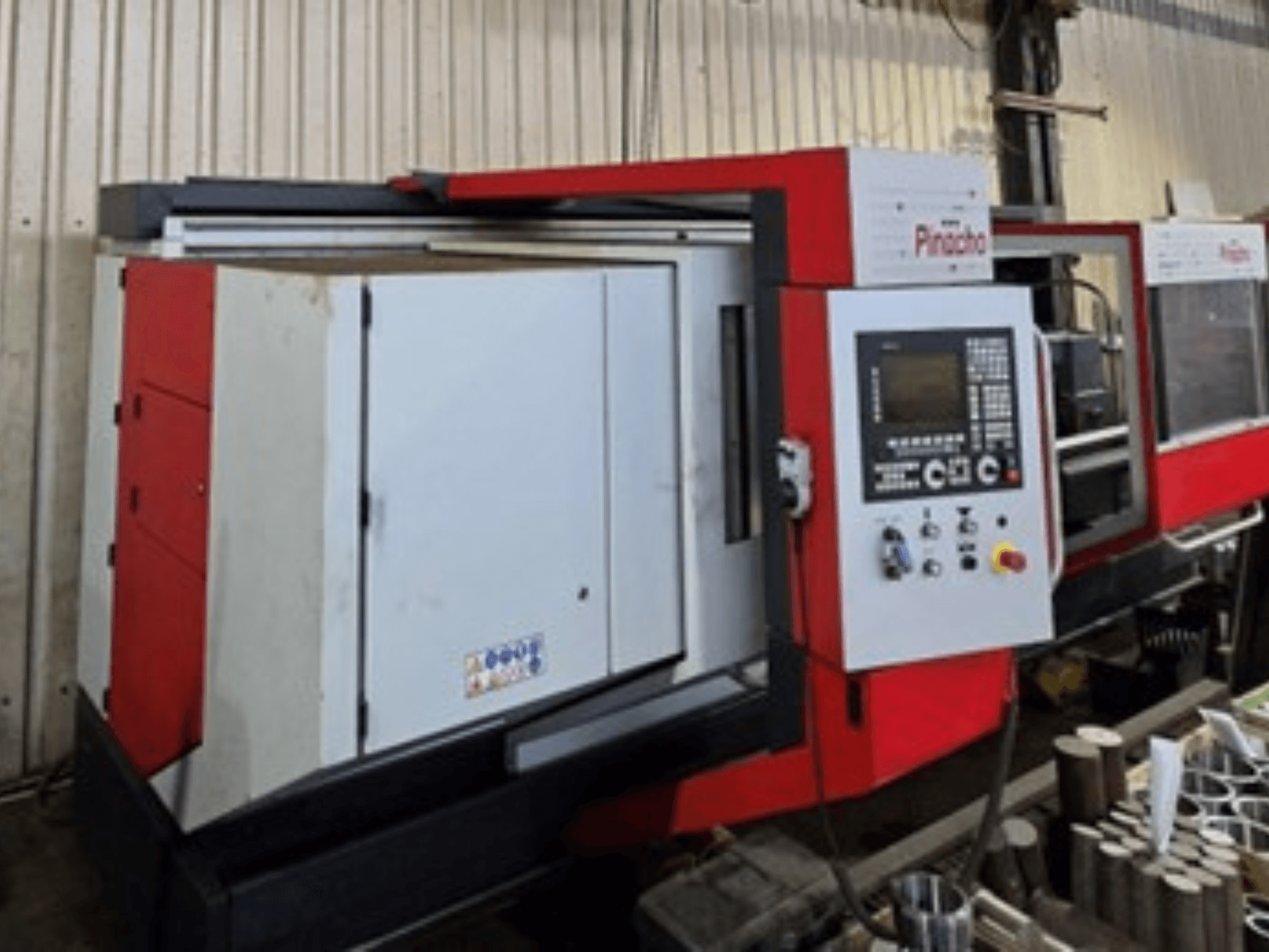 Machine CNC Pinacho en rouge et blanc, vue de côté, avec panneau de commande et matériaux au sol, montrant la configuration opérationnelle.