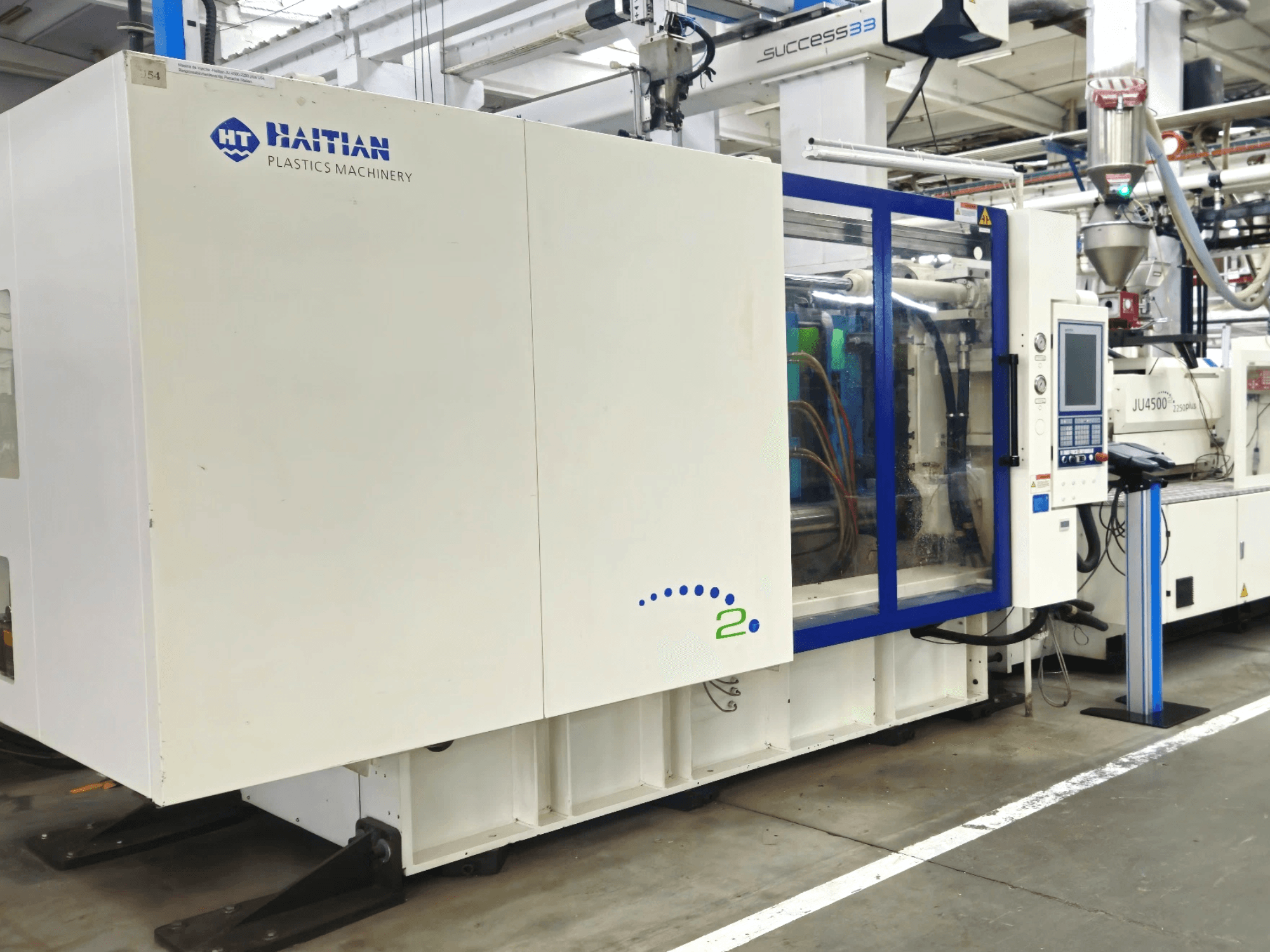 Modèle de Haitian Plastics Machinery, vue de face, montrant une machine avec un panneau de commande et des tuyaux visibles, dans un environnement industriel.