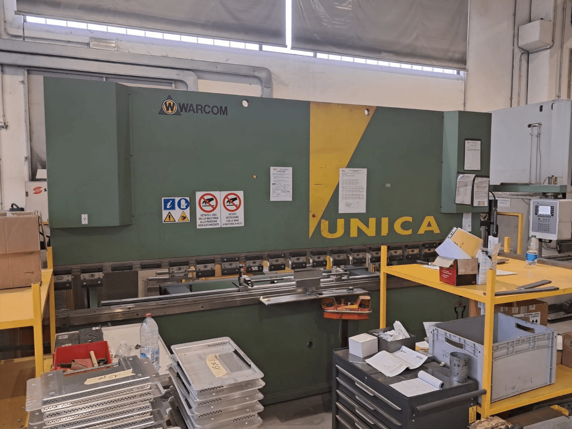 Presse hydraulique Warcom UNICA 100-40, vue de face, châssis vert rehaussé de touches de jaune, panneaux de sécurité visibles, outils et matériaux posés sur la table.