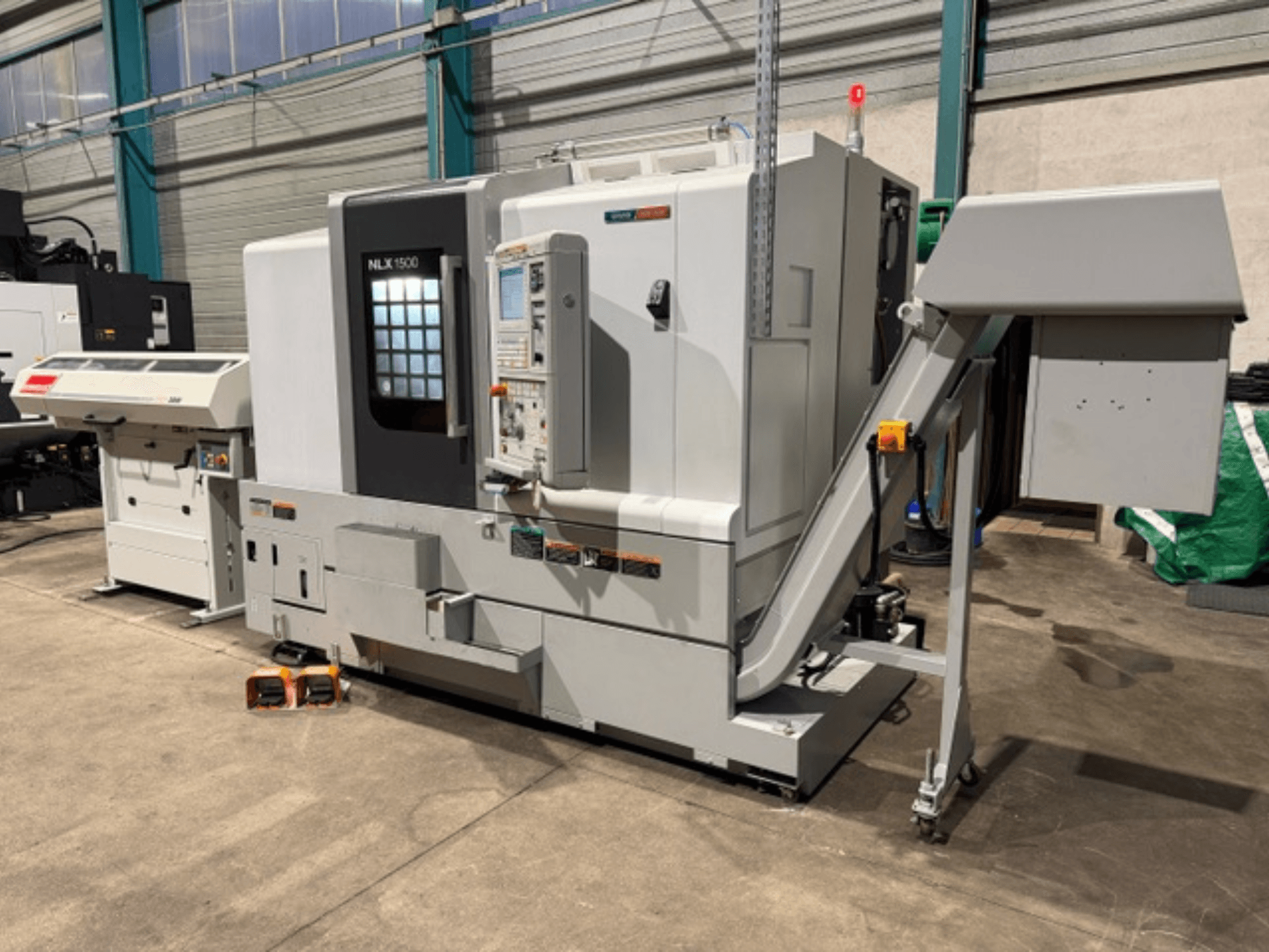 Machine CNC NLK 1500, vue de face, avec panneau de commande et trémie d'accessoires sur le côté, reposant sur un sol en béton.