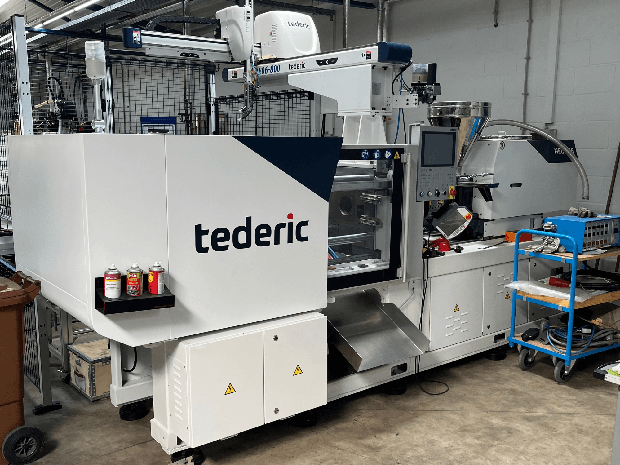 Presse à injection hydraulique TEDERIC NEO.E55/e110h dans un atelier, vue de profil avec le panneau de commande et les outils adjacents.