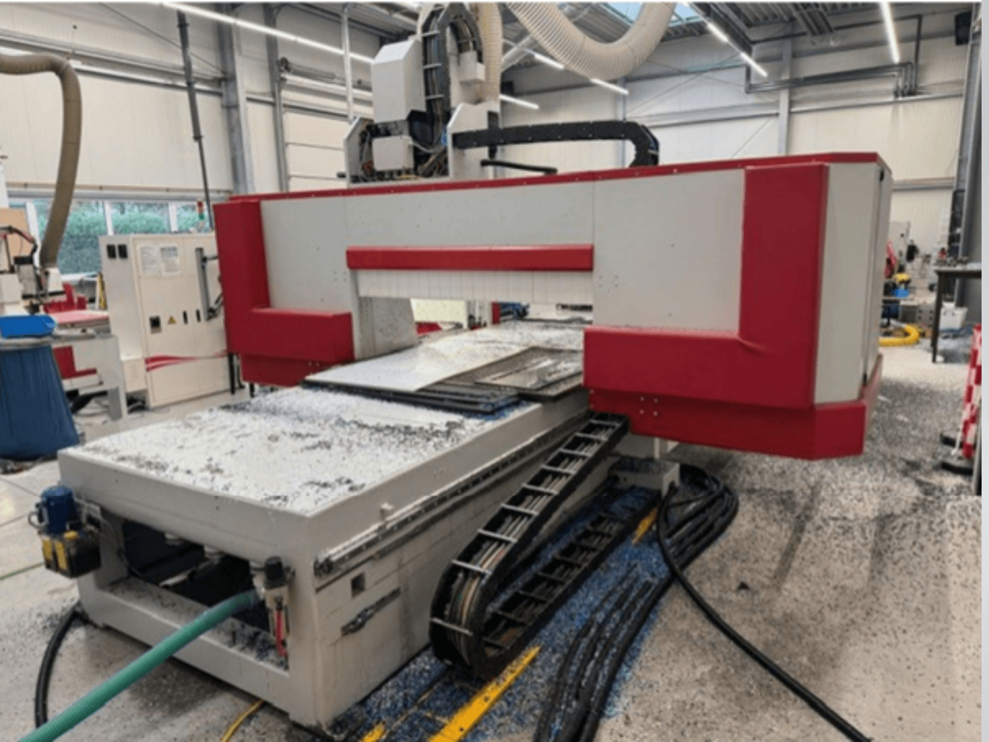 Machine CNC rouge et grise dans un atelier, vue latérale montrant la table des matériaux, le tuyau de refroidissement et la sciure de bois sur le sol.