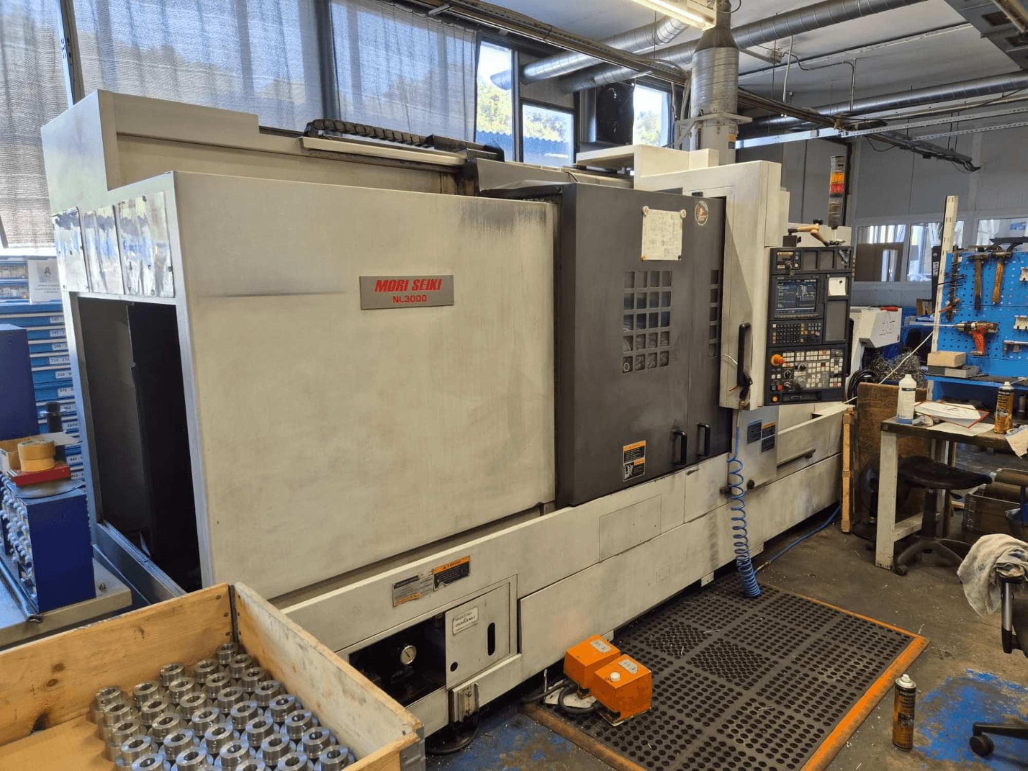 Tour CNC Mori Seiki NL3000 dans un atelier, vue latérale montrant le panneau de commande, les outils et les pièces métalliques dans une boîte en bois.