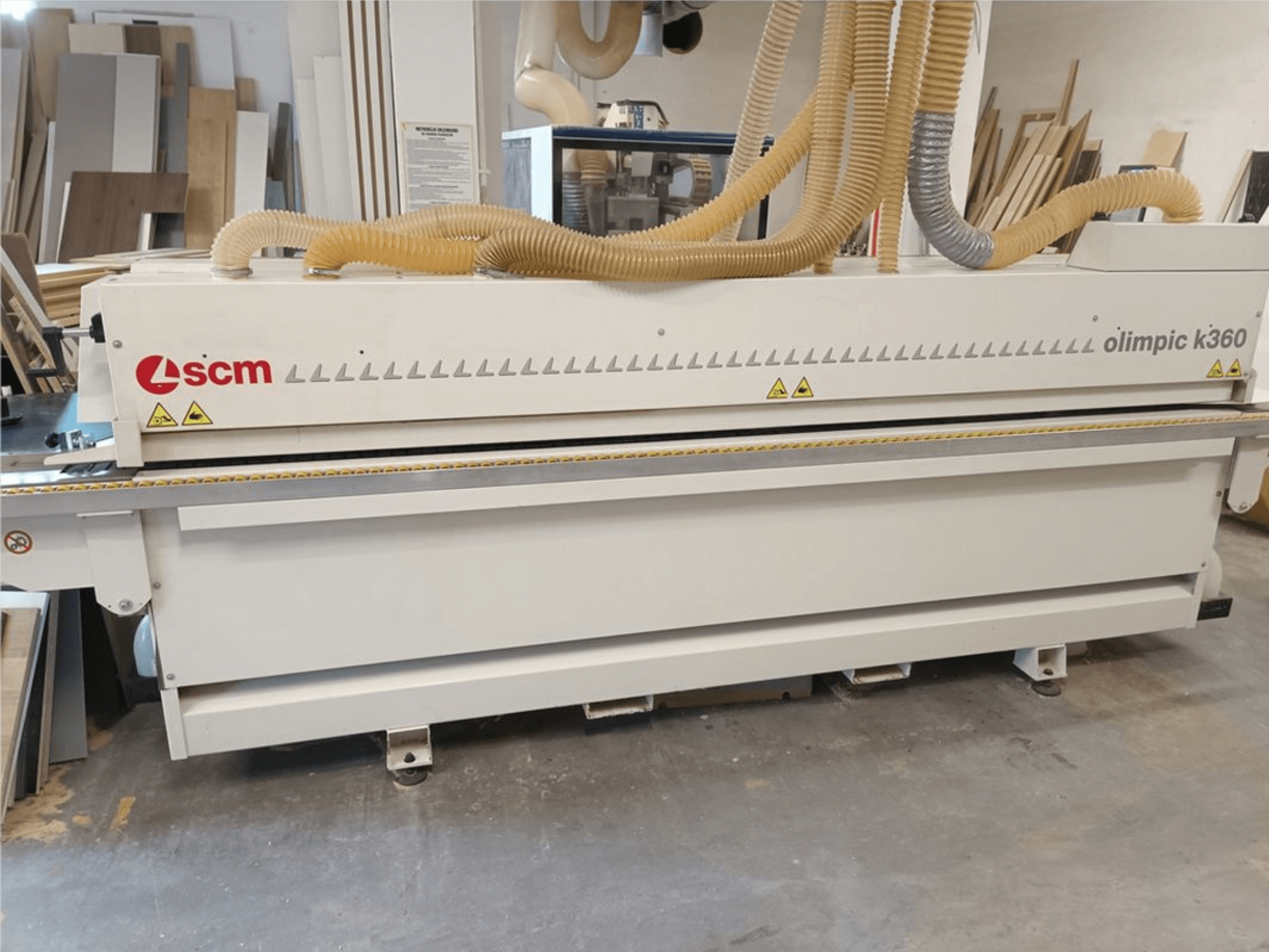 Plaqueuse de chants SCM Olimpic K360 T-ER1 dans un atelier, vue de face montrant les tuyaux d'aspiration de poussière et les étiquettes d'avertissement.