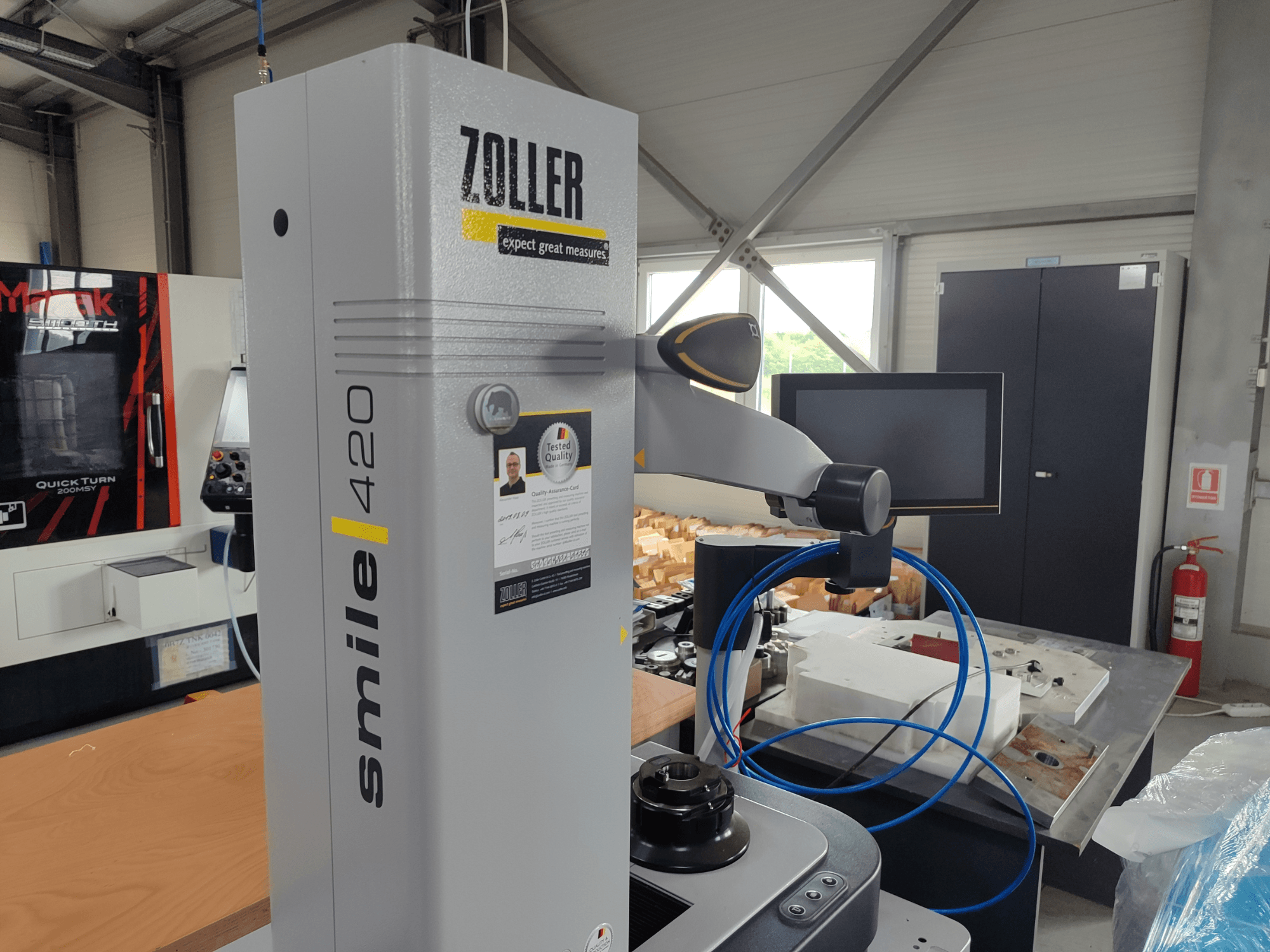 Appareil de mesure ZOLLER smile 420 avec moniteur, vue latérale dans un atelier, avec des tuyaux bleus et des machines en arrière-plan.