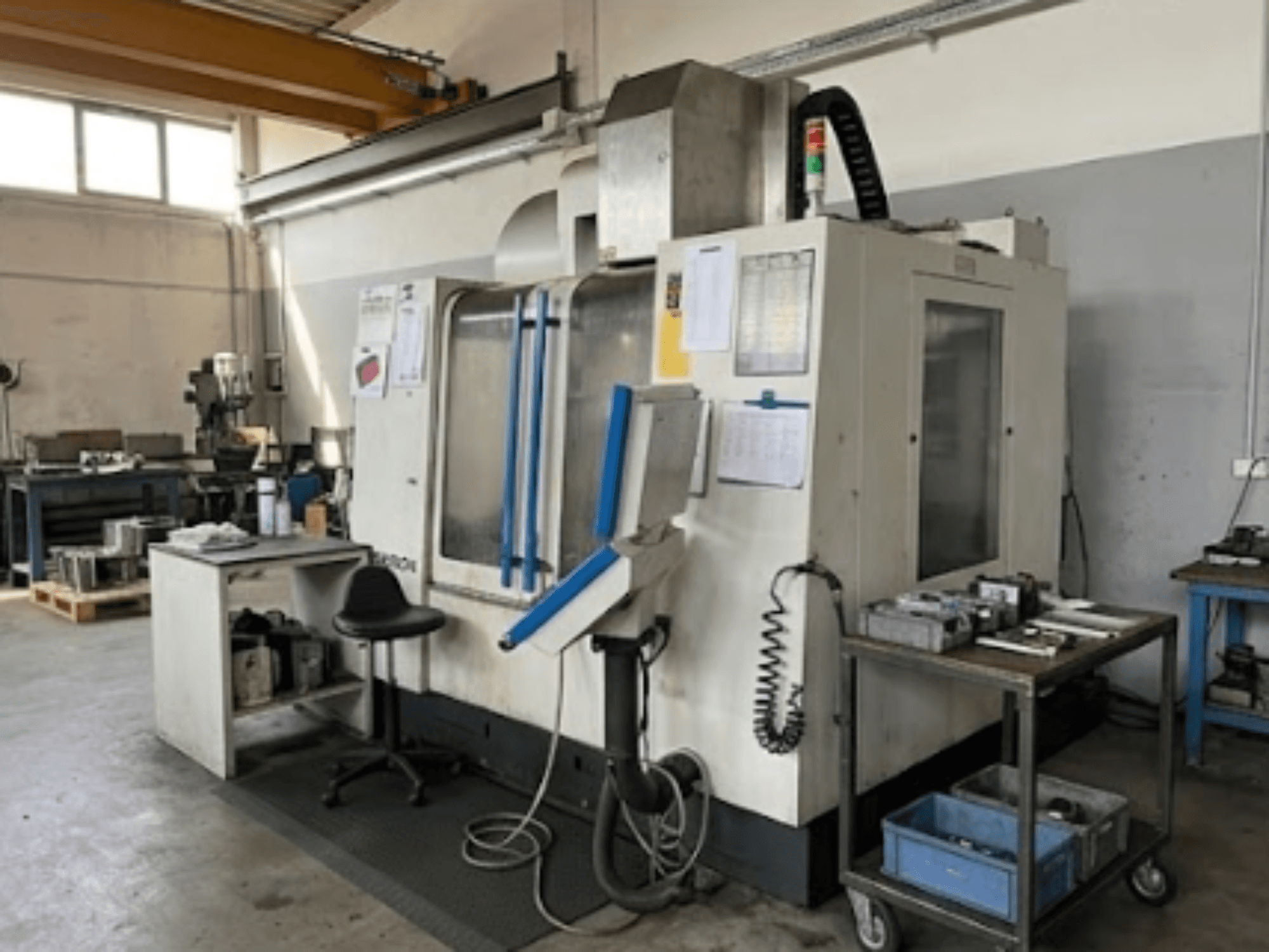 Centre d'usinage vertical MIKRON VCE 1000 Pro dans un atelier, vue de face avec des touches de bleu et une table de travail adjacente.