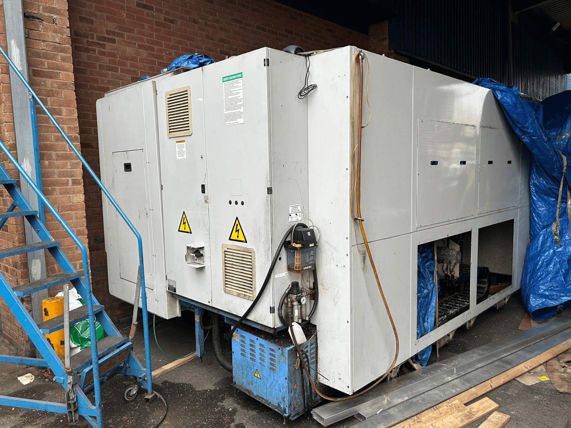 Vue latérale d'un grand refroidisseur industriel, dont la marque et le modèle ne sont pas clairs, montrant les connexions électriques, les panneaux d'avertissement et la bâche bleue.