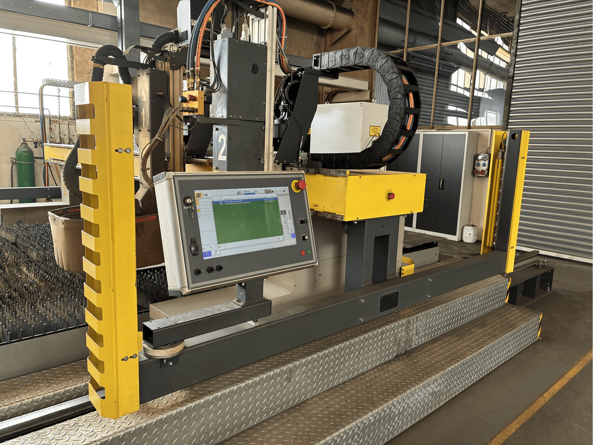 Machine à découper industrielle avec panneau de commande à écran tactile, protecteur de sécurité jaune, vue de face, base en caillebotis métallique visible.