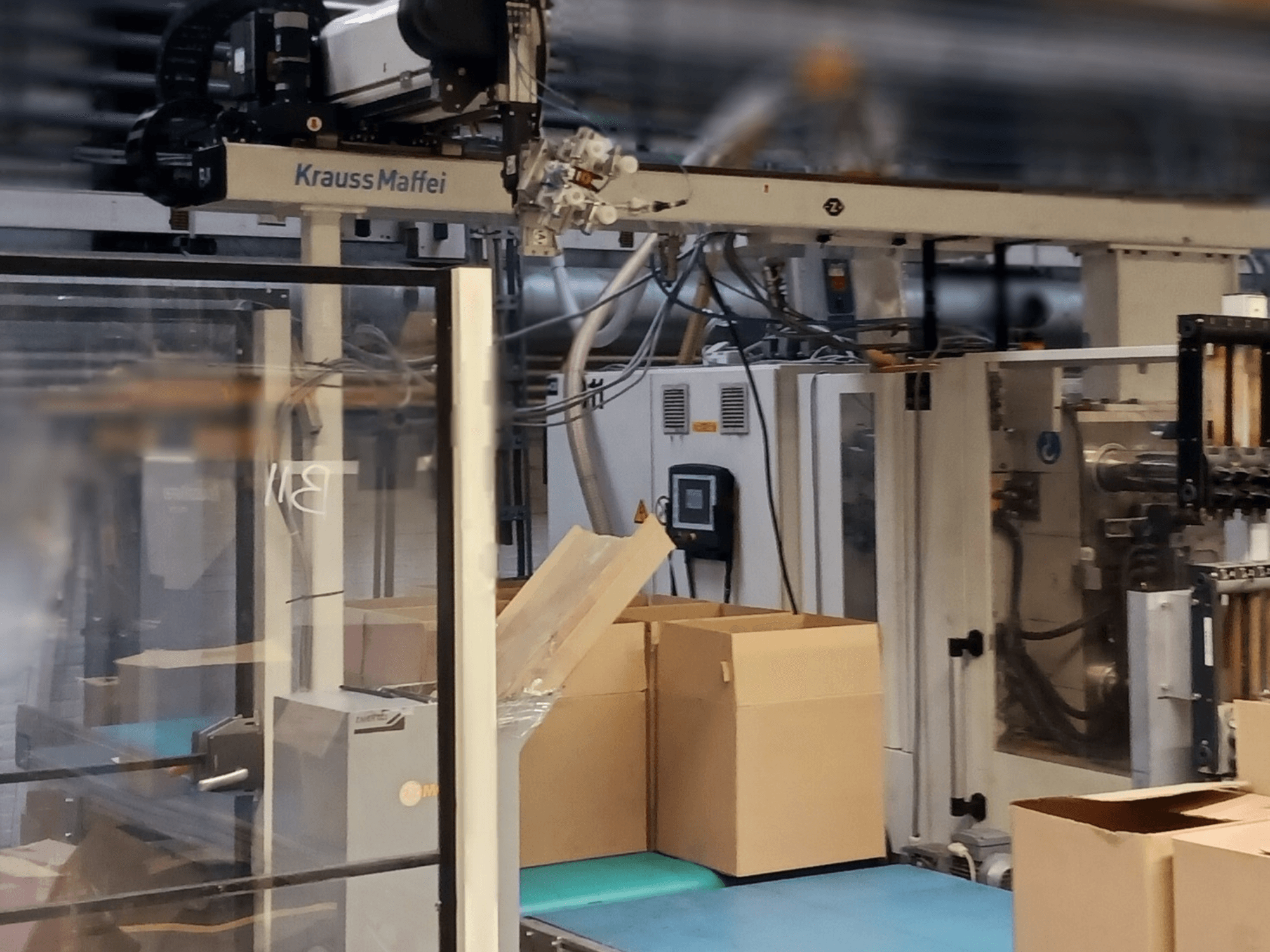 Système d'automatisation Krauss Maffei dans une usine, vue de face montrant des boîtes sur un convoyeur et des composants de machines en fonctionnement.