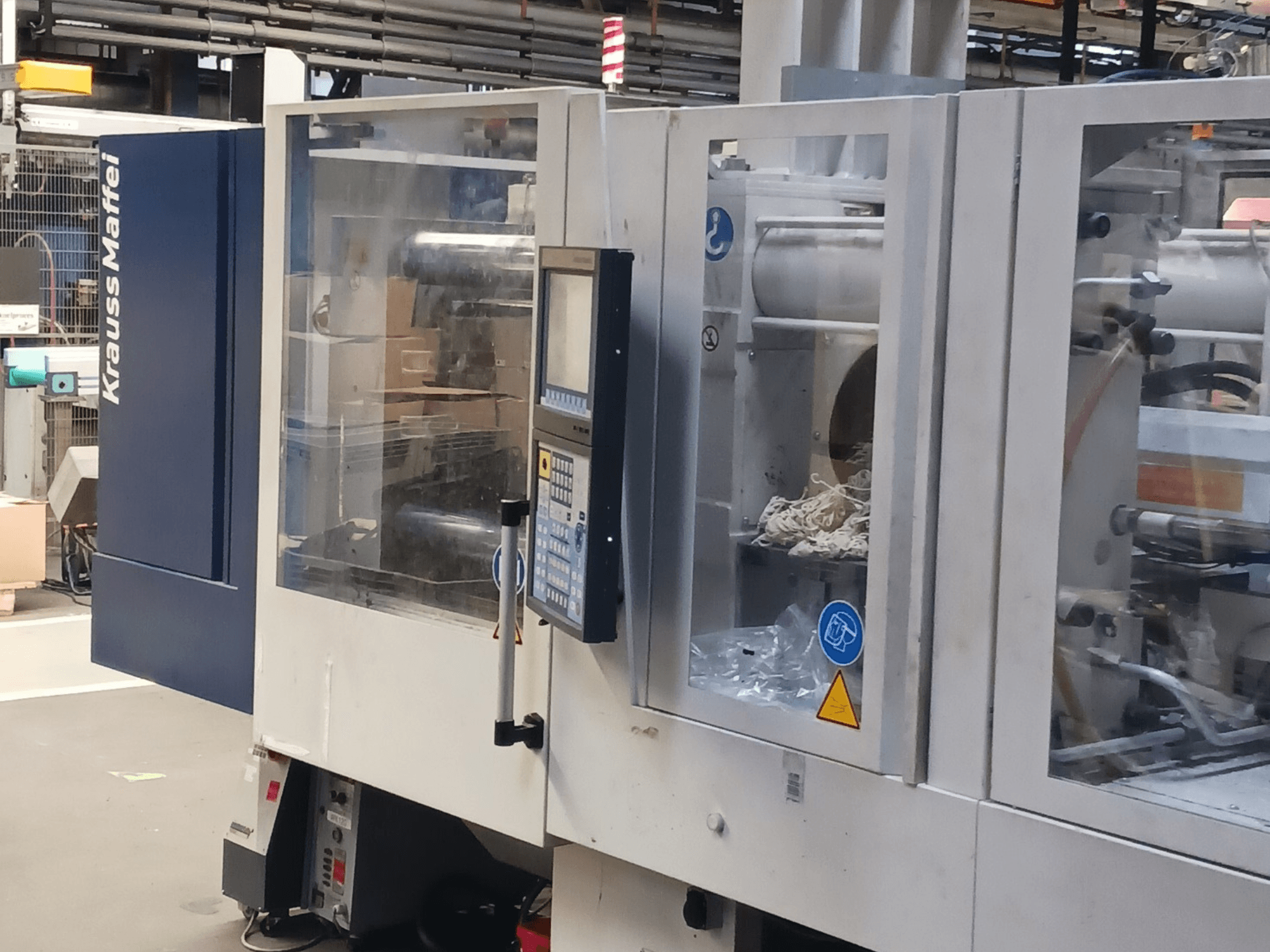 Machine de moulage par injection KraussMaffei, vue de côté, montrant le panneau de commande, les machines et les matériaux d'emballage à l'arrière-plan.