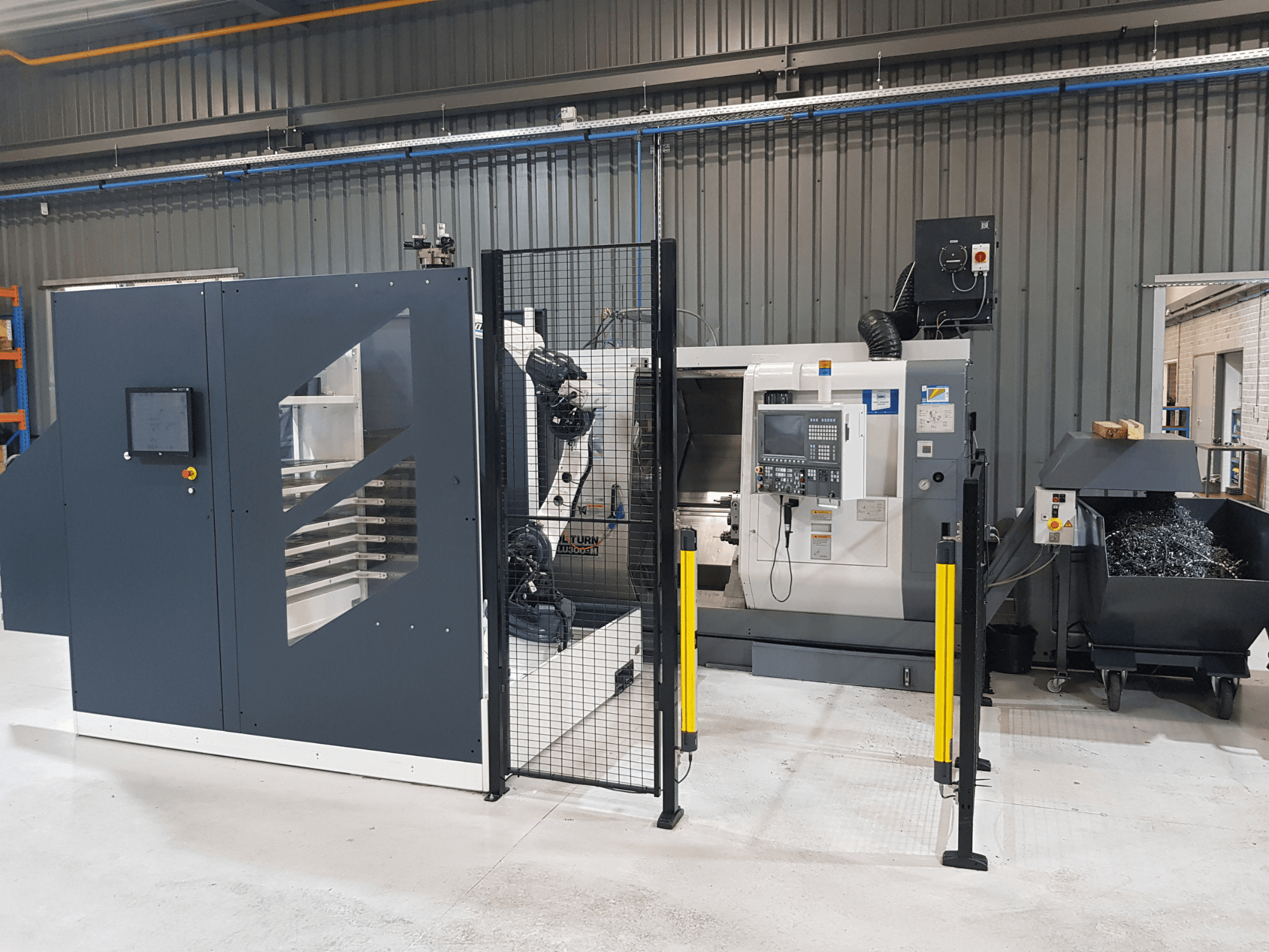 Tour automatisé et machine CNC ALTURN présentés dans une usine ; vue de face avec le panneau de commande et les barrières de sécurité visibles.