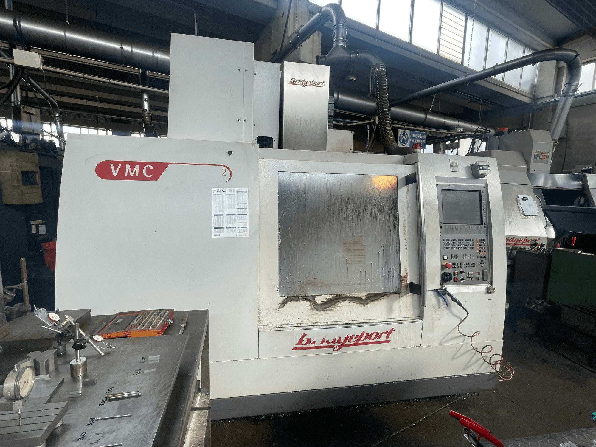 Centre d'usinage vertical Bridgeport VCM 800-30 dans un atelier, avec son panneau de commande et sa table porte-outils. Vue de face montrant les détails.