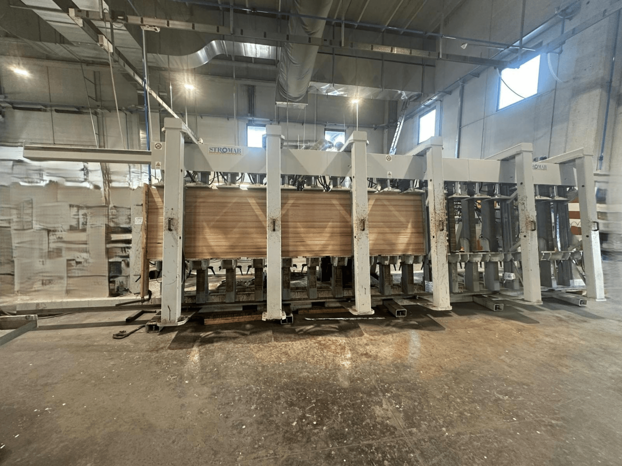 Machine industrielle STROMAB dans un entrepôt, vue de face, avec des panneaux de bois en cours de traitement, montrant la structure métallique et les pièces mécaniques.
