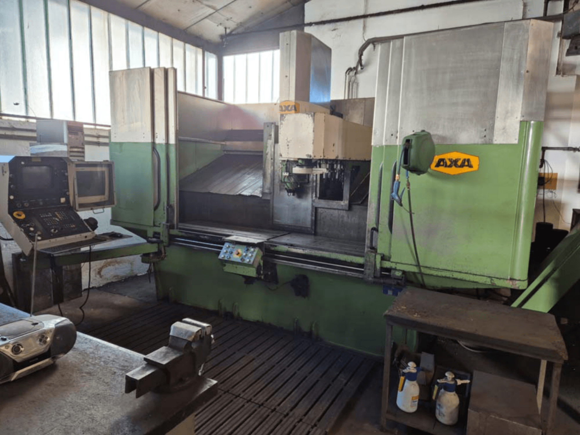 Centre d'usinage vertical AXA VSC-2M, vue de face, comprenant le panneau de commande et la zone de travail avec les outils de coupe et les produits de nettoyage.