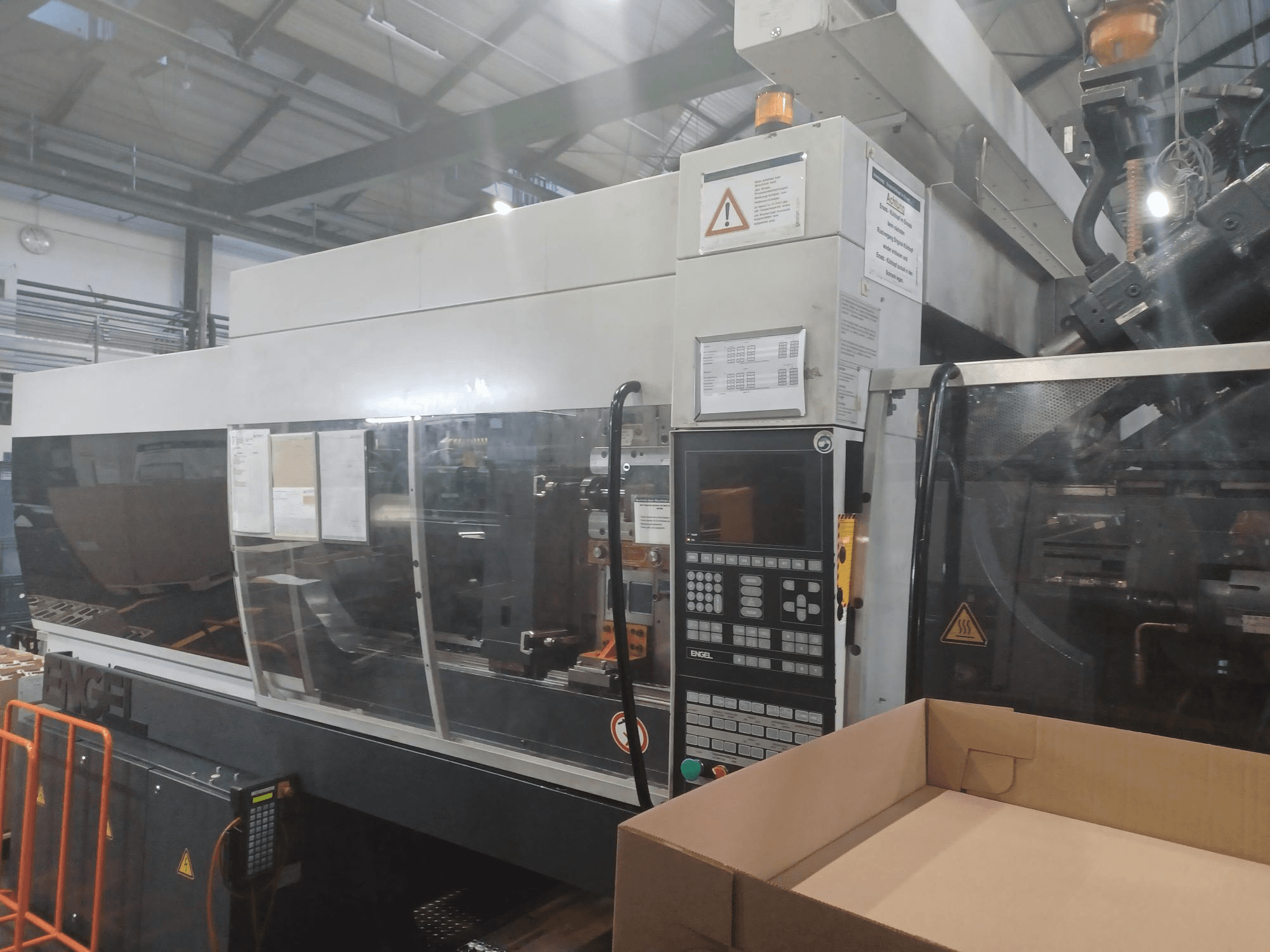 Machine de moulage par injection Engel, vue de face, avec le panneau de commande, les étiquettes de sécurité et une boîte vide placée à proximité.