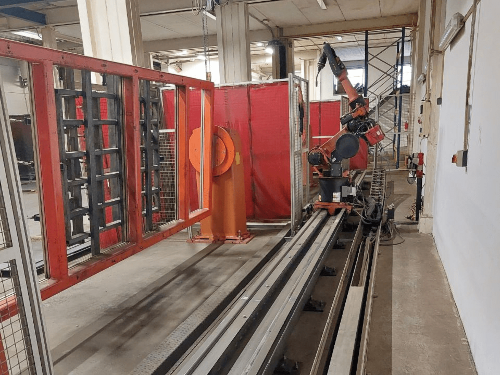 Bras robotique industriel aux couleurs orange et noire positionné sur des rails métalliques, entouré de barrières de protection rouges, vue de face.